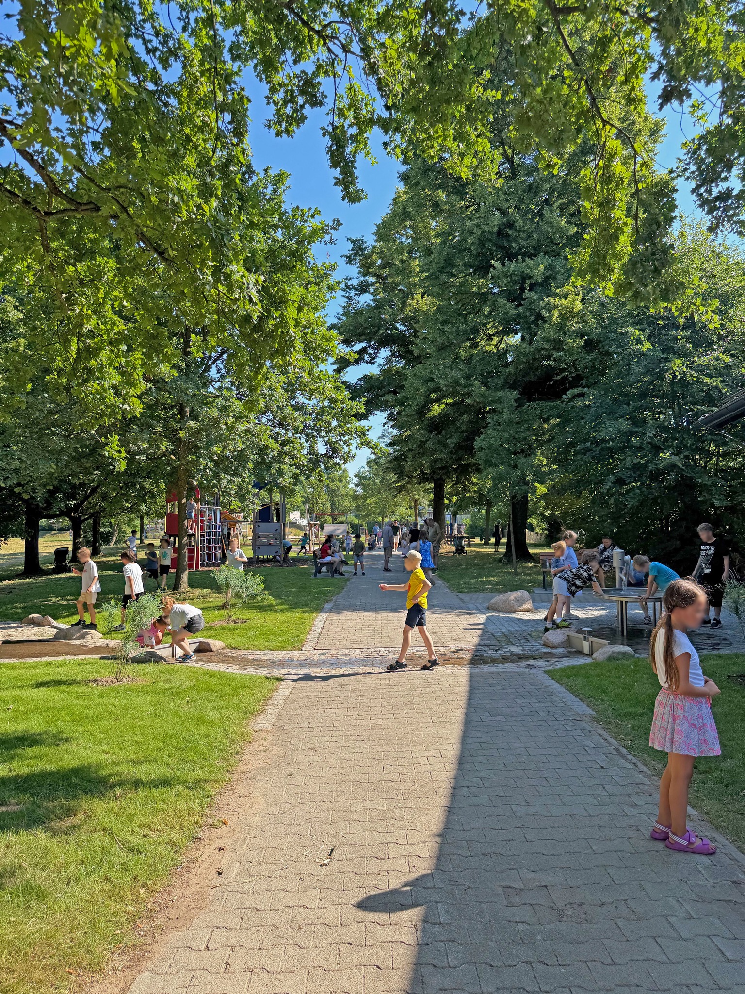 Ein gepflasterter Weg führt durch einen sonnigen Park, der von großen Bäumen gesäumt ist. Zahlreiche Kinder spielen verteilt im Bild: Einige nutzen rechts einen Wasserspielplatz, andere sind links an einem kleinen Wasserlauf zu sehen und eines läuft in der Mitte über den Weg. Im Hintergrund befinden sich weitere Personen und Spielgeräte. (KI-Alternativtext)