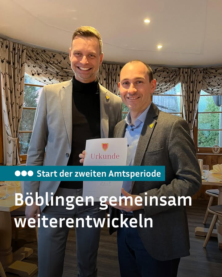 Dieser Alternativtext wurde KI-generiert: Zwei Personen stehen lächelnd nebeneinander, beide tragen Jacken und Hemden. Im Hintergrund ist ein Fenster mit Blick auf eine Landschaft zu sehen und ein Schild mit Text.