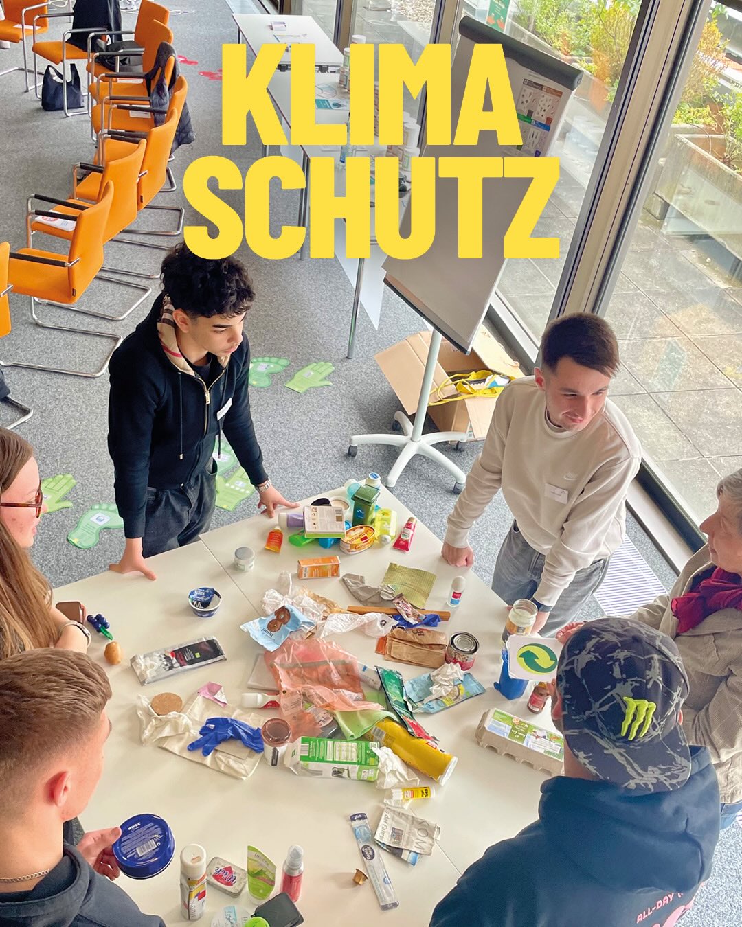 Mehrere Personen stehen um einen Tisch mit diversen Verpackungsabfällen und diskutieren. Im Hintergrund sind orangefarbene Stühle und ein Flipchart zu sehen. Große gelbe Schrift „KLIMASCHUTZ“ überlagert das Bild. (KI-Alternativtext)