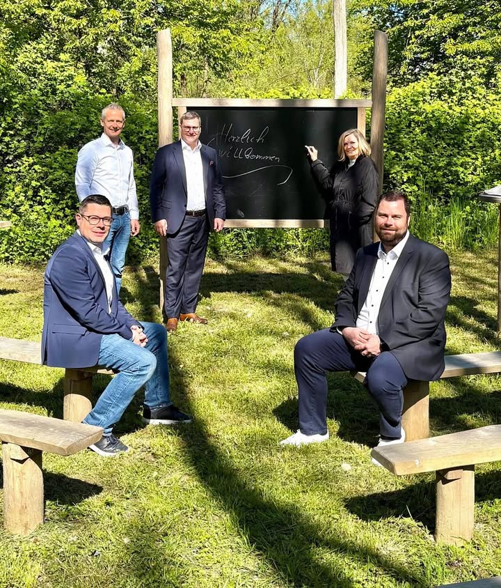 Dieser Alternativtext wurde KI-generiert: Mehrere Personen in formeller Kleidung stehen und sitzen vor einer grünen Hecke und einer Schultafel. Das Bild ist farbig und im Freien aufgenommen.