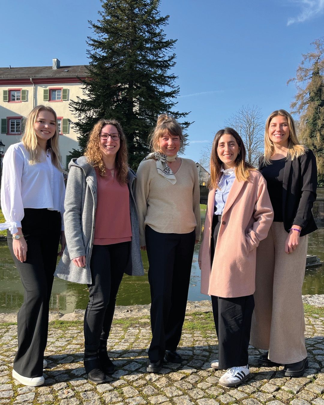 Dieser Alternativtext wurde KI-generiert: Eine Gruppe von Personen steht im Freien vor einer Gebäudezeile und Bäumen. Im Hintergrund befindet sich ein Teich oder ein kleiner See.