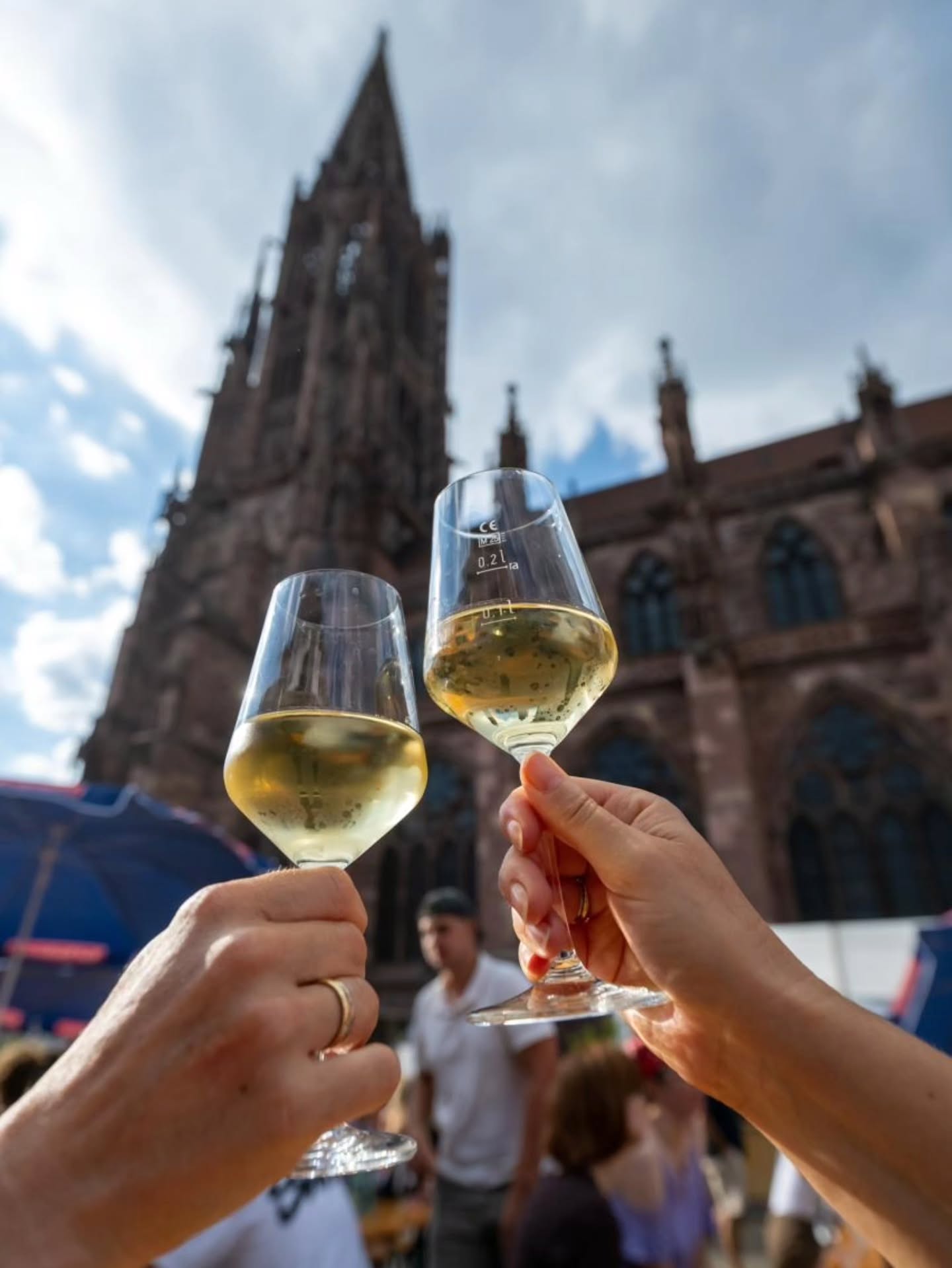 Zwei Hände halten Weingläser mit Weißwein und stoßen an. Im Hintergrund erhebt sich der hohe Turm einer gotischen Kathedrale vor einem blauen Himmel. (KI-Alternativtext)