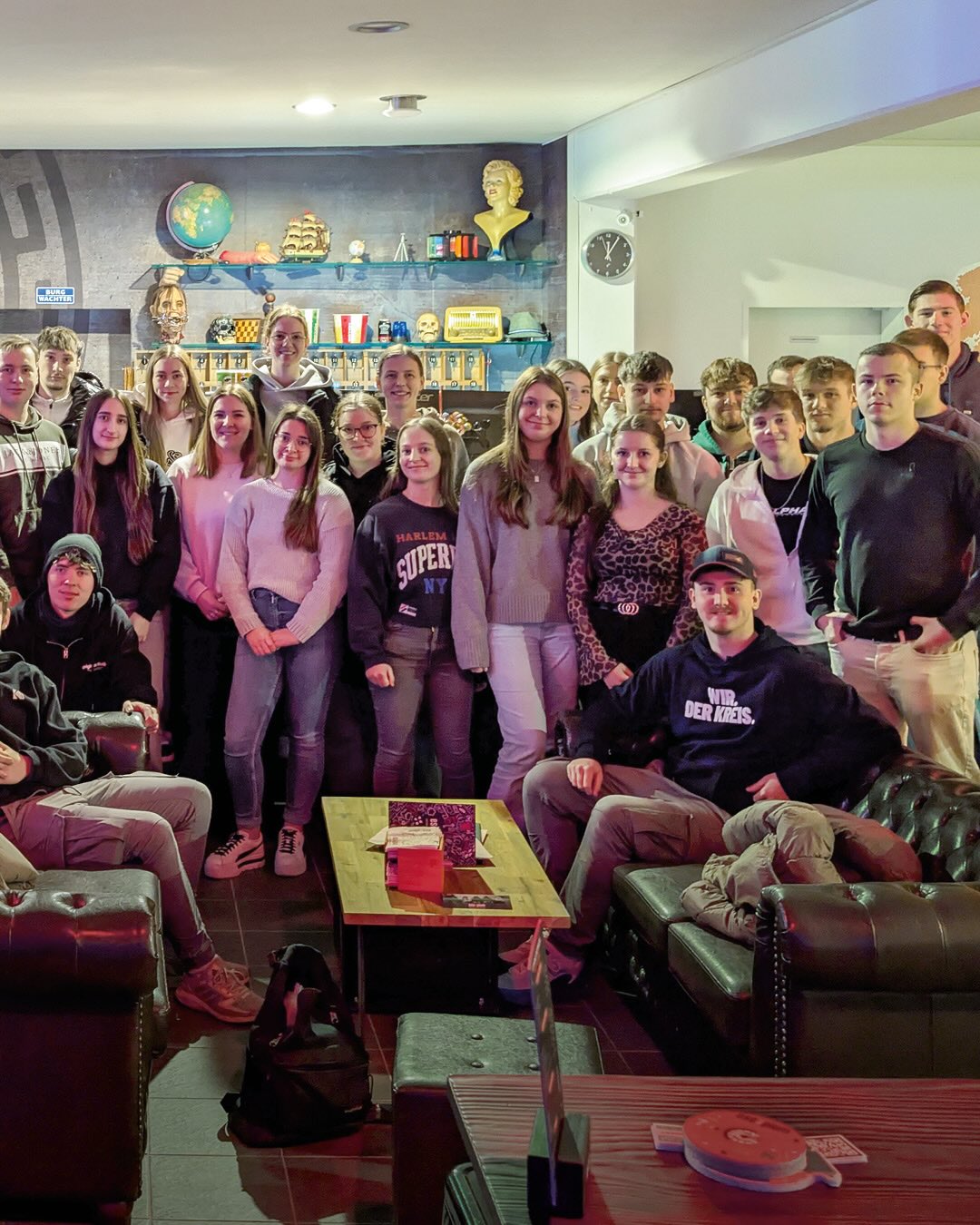 Dieser Alternativtext wurde KI-generiert: Eine Gruppe von Personen posiert in einem Raum mit Sofas und Dekorationen an der Wand. Im Hintergrund befindet sich eine Sammlung von Objekten auf einem Regal.