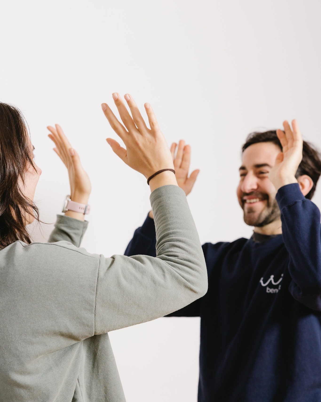 Zwei Personen geben sich mit erhobenen Händen einen High-Five vor weißem Hintergrund. Eine trägt ein graues Oberteil und eine Armbanduhr, die andere ein dunkelblaues Sweatshirt mit kleinem Logo. Beide lächeln. (KI-Alternativtext)