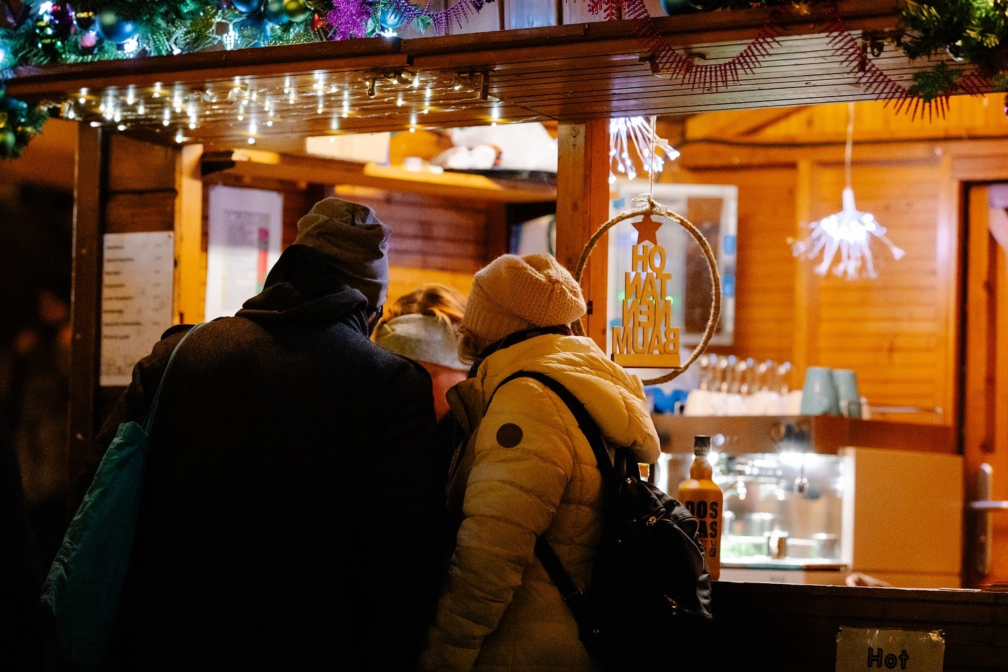 Zwei Personen stehen an einem Weihnachtsmarktstand und scheinen etwas zu bestellen. Der Stand ist beleuchtet und weihnachtlich geschmückt.