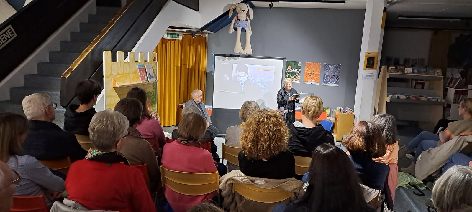 Dieser Alternativtext wurde KI-generiert: Eine Gruppe von Personen sitzt auf Stühlen und schaut auf eine Projektion an der Wand. Im Hintergrund ist eine Treppe und ein Raum mit Regalen zu sehen.