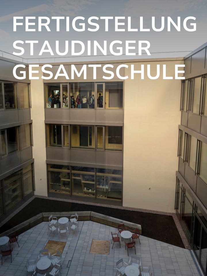 Ein modernes Schulgebäude mit einem gepflasterten Innenhof, in dem Tische und Stühle stehen. In den Fenstern des zweiten Obergeschosses blicken mehrere Personen nach draußen. Oben im Bild ist in großen weißen Buchstaben der Text „FERTIGSTELLUNG STAUDINGER GESAMTSCHULE“ eingeblendet. (KI-Alternativtext)