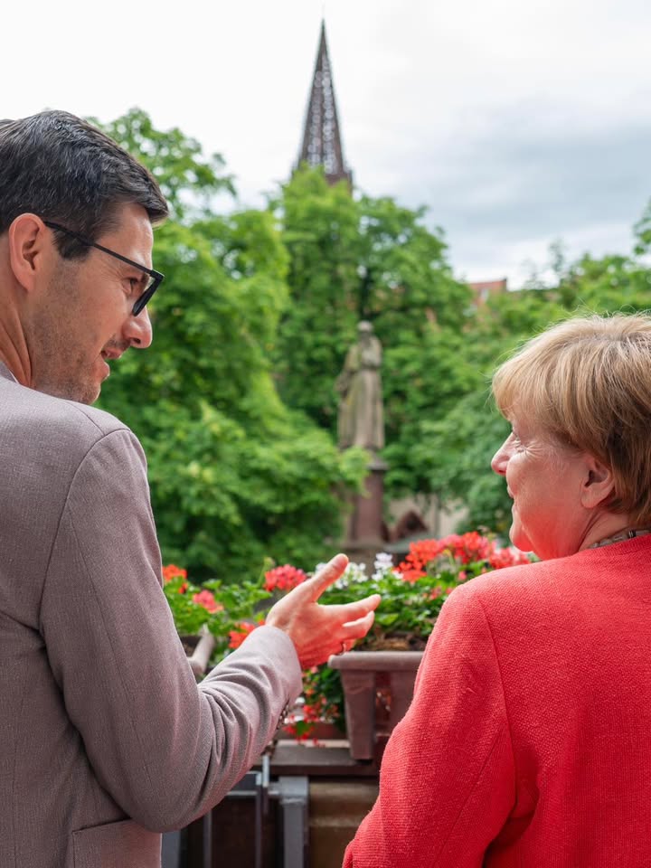 Zwei Personen unterhalten sich im Freien, eine gestikuliert mit der Hand. Im Hintergrund sind Blumen, Bäume, eine Statue und ein Kirchturm zu sehen. (KI-Alternativtext)