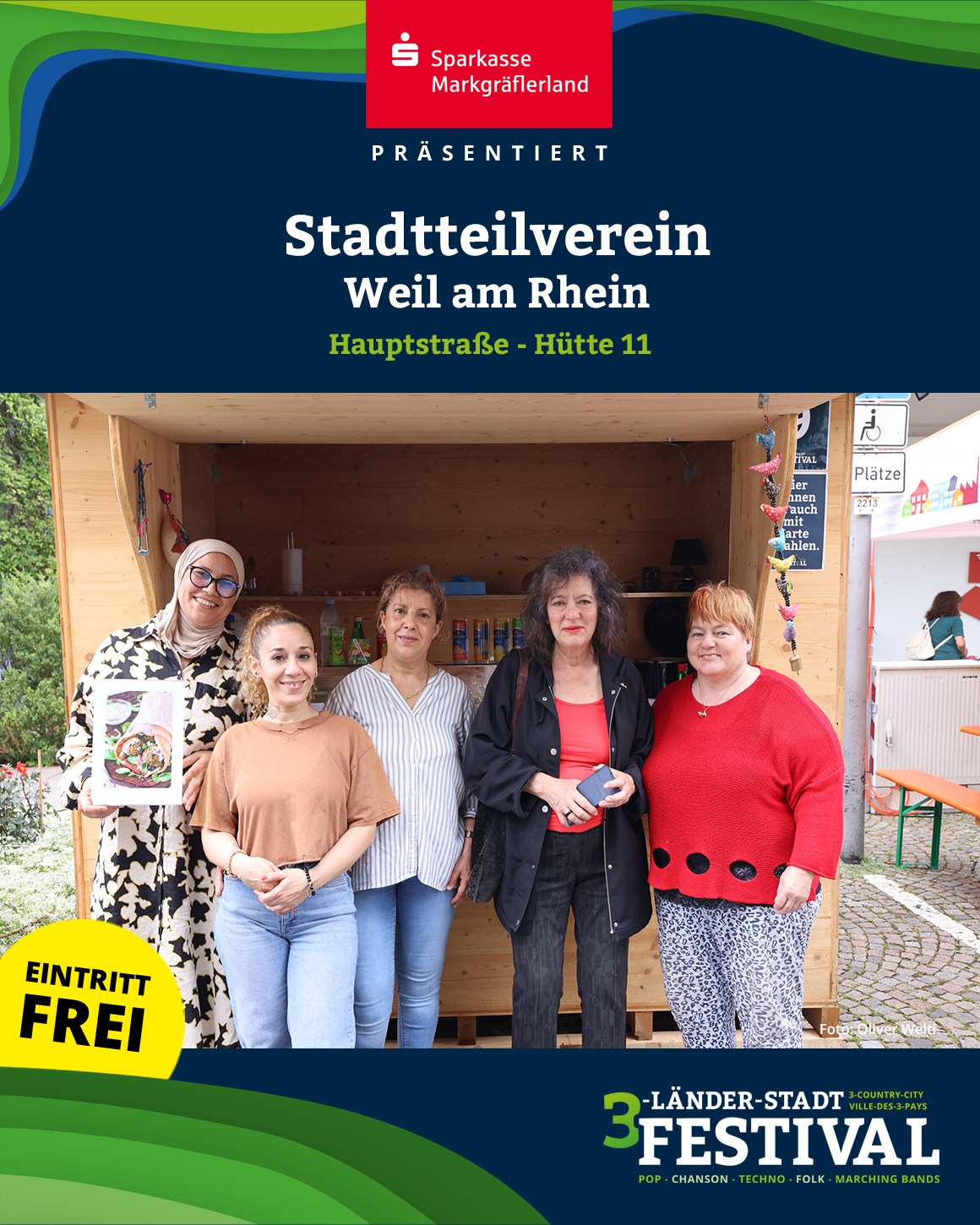 Eine Gruppe von Menschen steht vor einer Holzbude. Auf dem Bild sind auch Logos und Schriftzüge zu sehen, die auf eine Veranstaltung hinweisen.