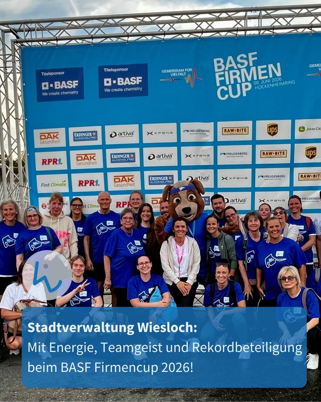 Gruppe von Menschen in blauen T-Shirts posiert vor einem blauen Banner mit Sponsorenlogos und der Aufschrift „BASF Firmencup 09. Juni 2026 Hockenheimring“. Eine Person im Hundemaskottchen steht in der Mitte, unten ist ein Textfeld mit „Stadtverwaltung Wiesloch: Mit Energie, Teamgeist und Rekordbeteiligung beim BASF Firmencup 2026!“. (KI-Alternativtext)