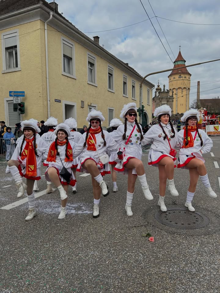 Dieser Alternativtext wurde KI-generiert: Eine Gruppe von Personen in traditioneller Tracht tanzt auf einer Straße vor einer Gebäudezeile. Im Hintergrund ist ein Turm mit einer roten Spitze zu sehen.