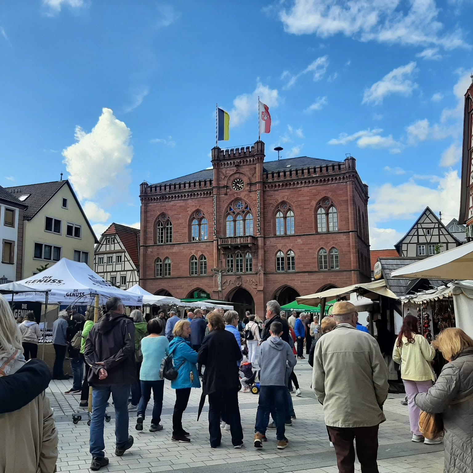 Auf einem Marktplatz steht ein großes Gebäude mit vielen Menschen davor. Es wehen zwei Flaggen an einem Fahnenmast auf dem Gebäude.