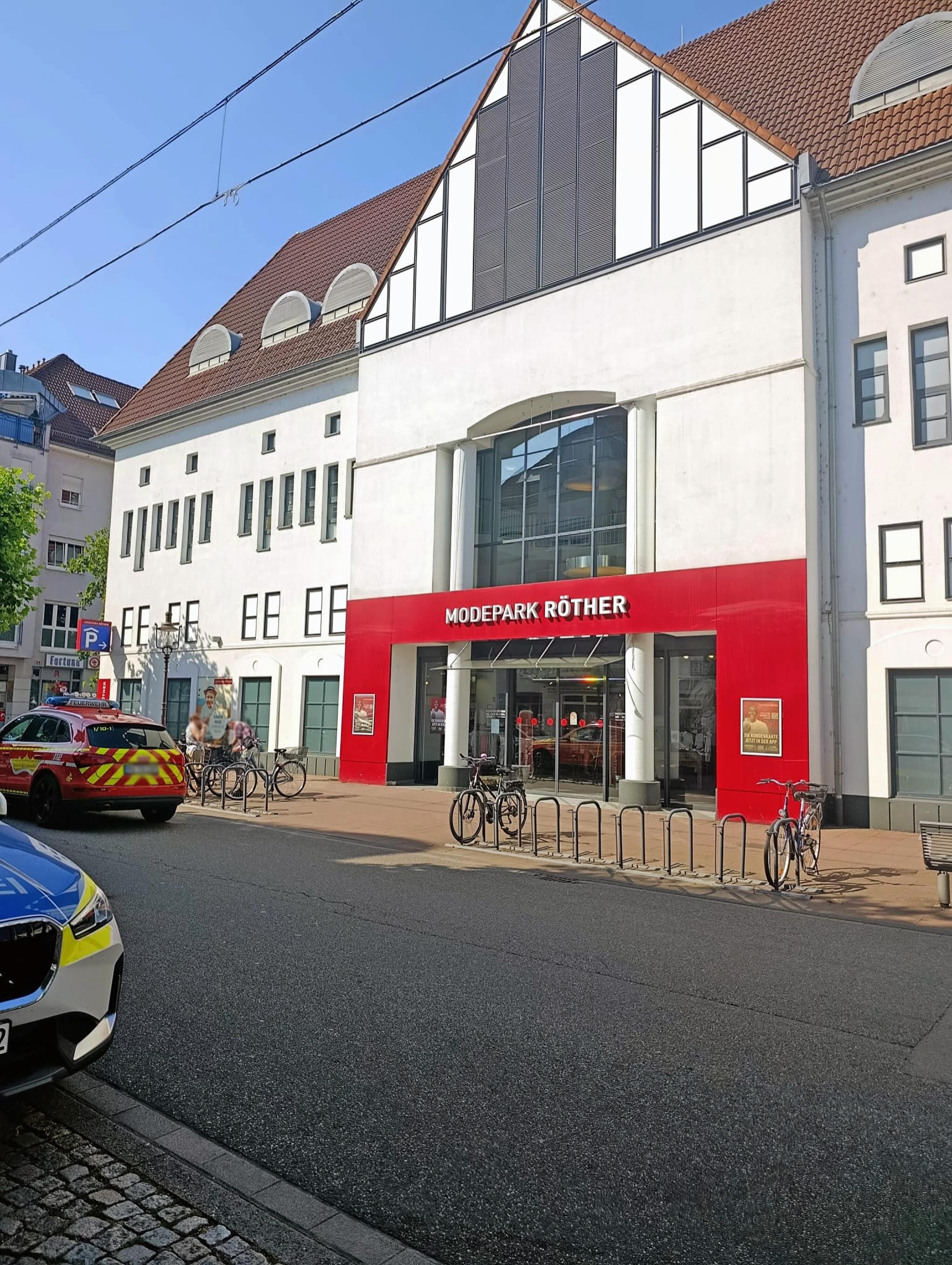 Ein weißes Gebäude mit rotem Ladeneingang und der Aufschrift "MODEPARK RÖTHER" steht an einer Straße, vor dem mehrere Fahrräder abgestellt sind. Links im Bild parkt ein rotes Feuerwehrfahrzeug, neben dem Personen stehen, und im Vordergrund ist der vordere Teil eines Polizeiwagens zu sehen. (KI-Alternativtext)