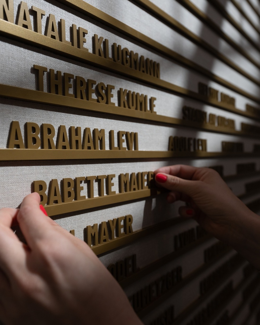 Zwei Hände befestigen ein goldenes Namensschild mit der Aufschrift "BABETTE MAIER" an einer Wand. Die Wand ist mit weiteren Reihen goldener Buchstaben beschriftet, darunter die Namen "NATALIE KLUGMANN", "THERESE KUMLE" und "ABRAHAM LEVI". (KI-Alternativtext)