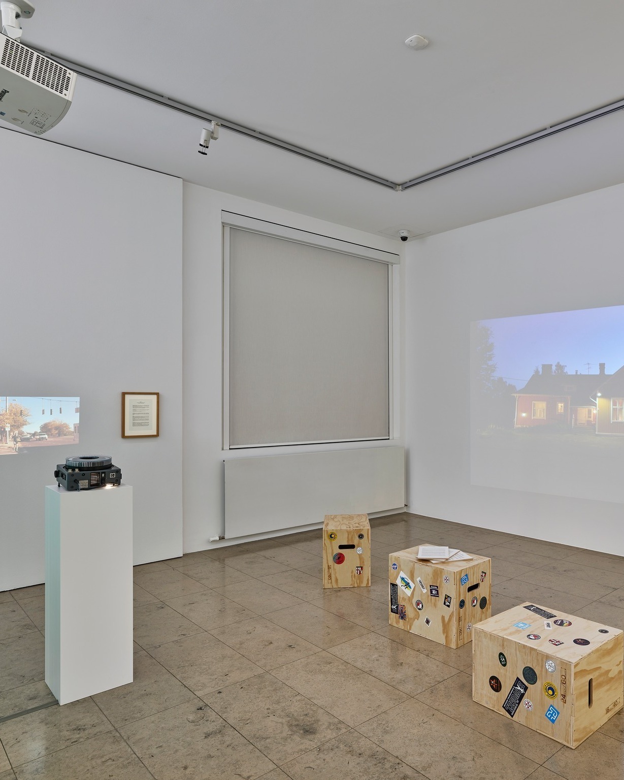 Ein heller Ausstellungsraum mit weißen Wänden und Fliesenboden. Links steht ein Projektor auf einem weißen Sockel, daneben ist eine kleine Straßenszene an die Wand projiziert sowie ein gerahmter Text. In der Mitte verdeckt ein Rollladen ein Fenster, während rechts eine große Projektion eines Hauses zu sehen ist. Auf dem Boden stehen drei mit Aufklebern versehene Holzkisten. (KI-Alternativtext)