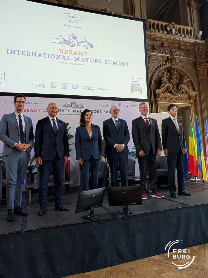Sechs Personen in Anzügen stehen auf einer Bühne vor einem großen Bildschirm mit der Aufschrift „URBAN7 INTERNATIONAL MAYORS SUMMIT“ und dem Datum „June 3–4, 2026“ in Nancy, Frankreich. Rechts im Hintergrund sind mehrere Flaggen und ein reich verzierter Bogen zu sehen. (KI-Alternativtext)