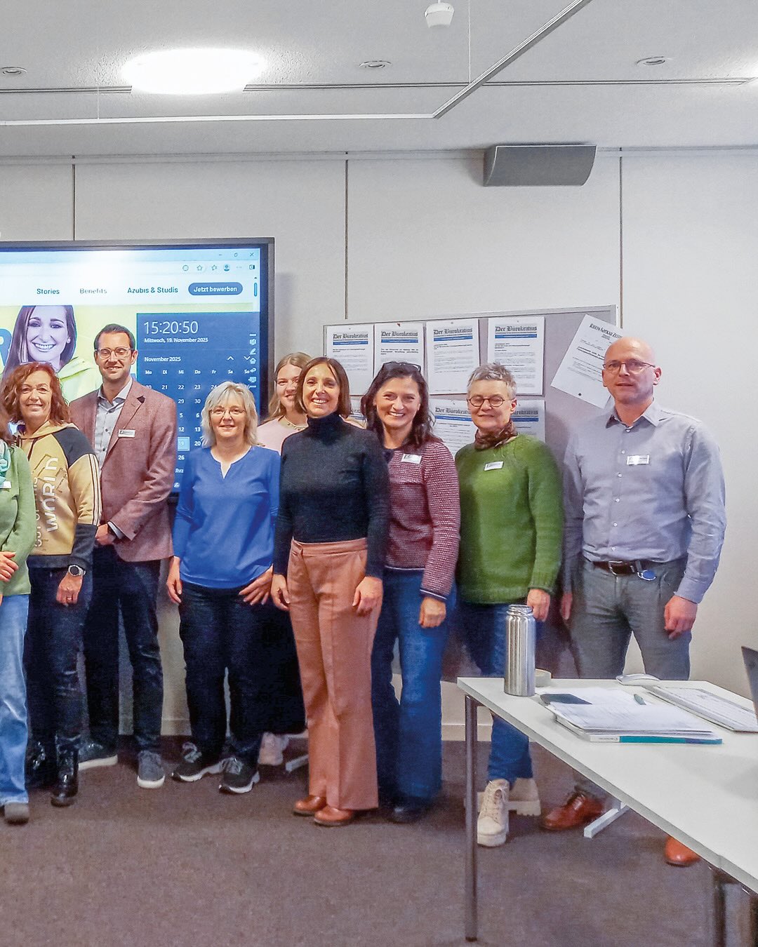 Dieser Alternativtext wurde KI-generiert: Eine Gruppe von Menschen steht in einem Innenraum vor einer Wand mit Postern und einem Bildschirm. Auf dem Bildschirm ist eine Person zu sehen und verschiedene Daten werden angezeigt.