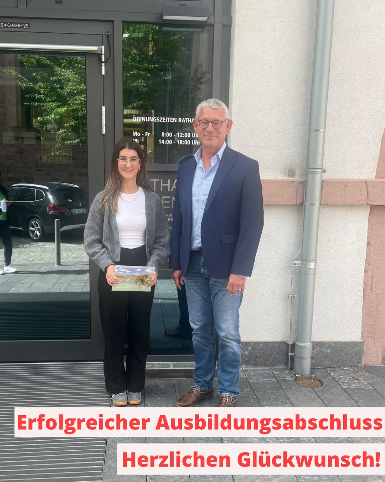 Zwei Personen stehen vor einem Gebäude, auf dem Öffnungszeiten stehen. Auf dem Foto ist der Text "Erfolgreicher Ausbildungsabschluss. Herzlichen Glückwunsch!" zu lesen.