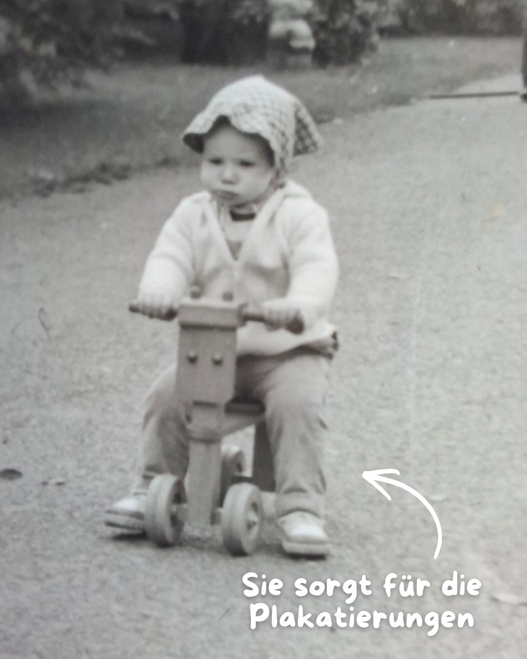 Dieser Alternativtext wurde KI-generiert: Schwarz-weißes Foto eines Kindes, das auf einem Dreirad sitzt. Das Kind trägt eine Kopfbedeckung und sieht in Richtung der Kamera.