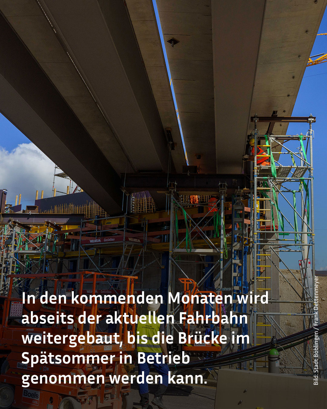 Unter einer Betonbrücke arbeiten Personen auf Gerüsten und mit Hebebühnen am Weiterbau; im Hintergrund ist blauer Himmel mit Wolken zu sehen. Ein Text überlagert das Bild und informiert über die geplante Inbetriebnahme im Spätsommer. (KI-Alternativtext)