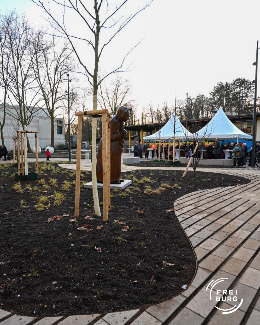 Dieser Alternativtext wurde KI-generiert: Ein gepflasterter Weg führt an einer Skulptur und mehreren Zelten vorbei. Im Hintergrund sind Gebäude und Bäume zu sehen.