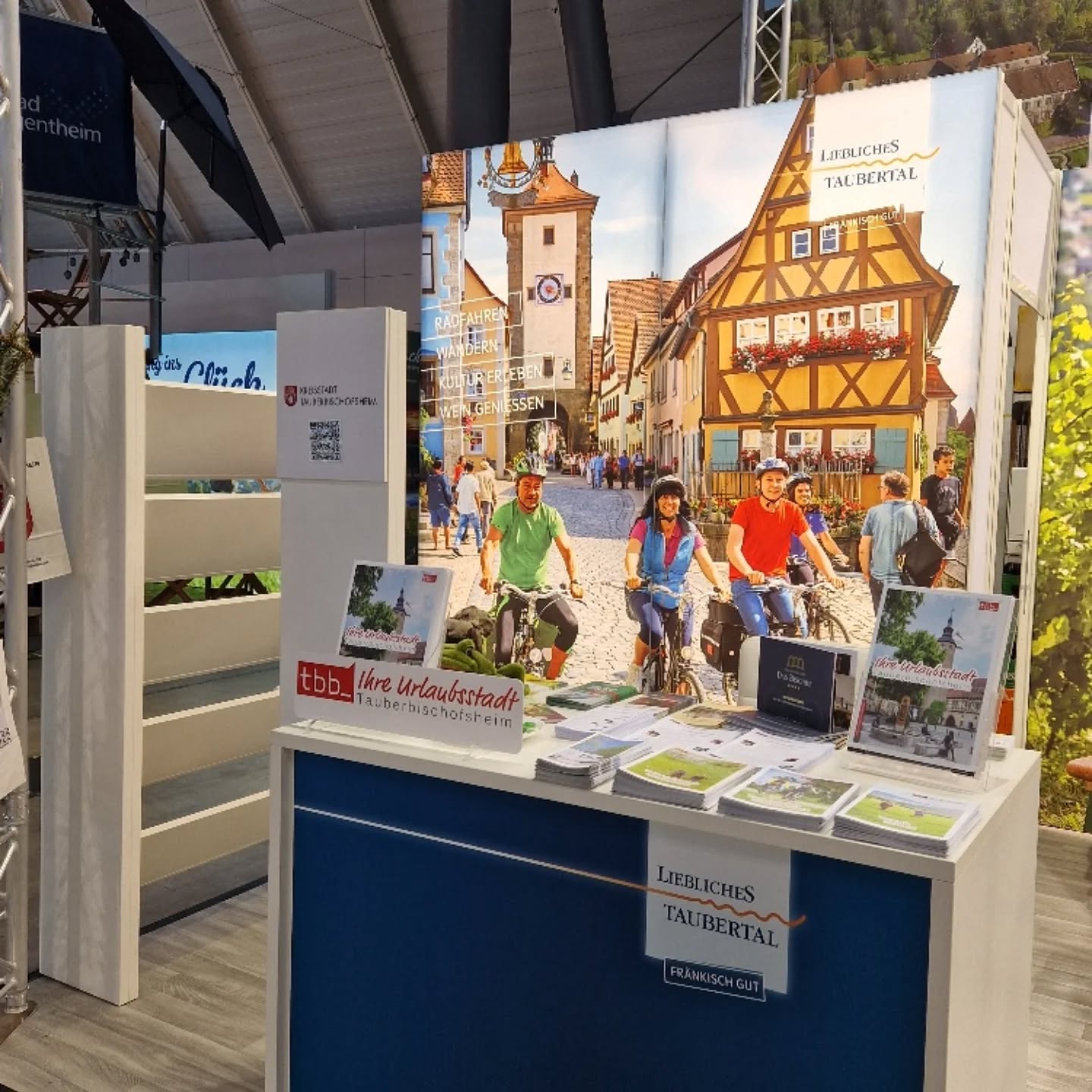 Ein Messestand wirbt für eine Urlaubsregion mit Bildern einer malerischen Stadt und Radfahrern. Broschüren und Infomaterial liegen bereit.