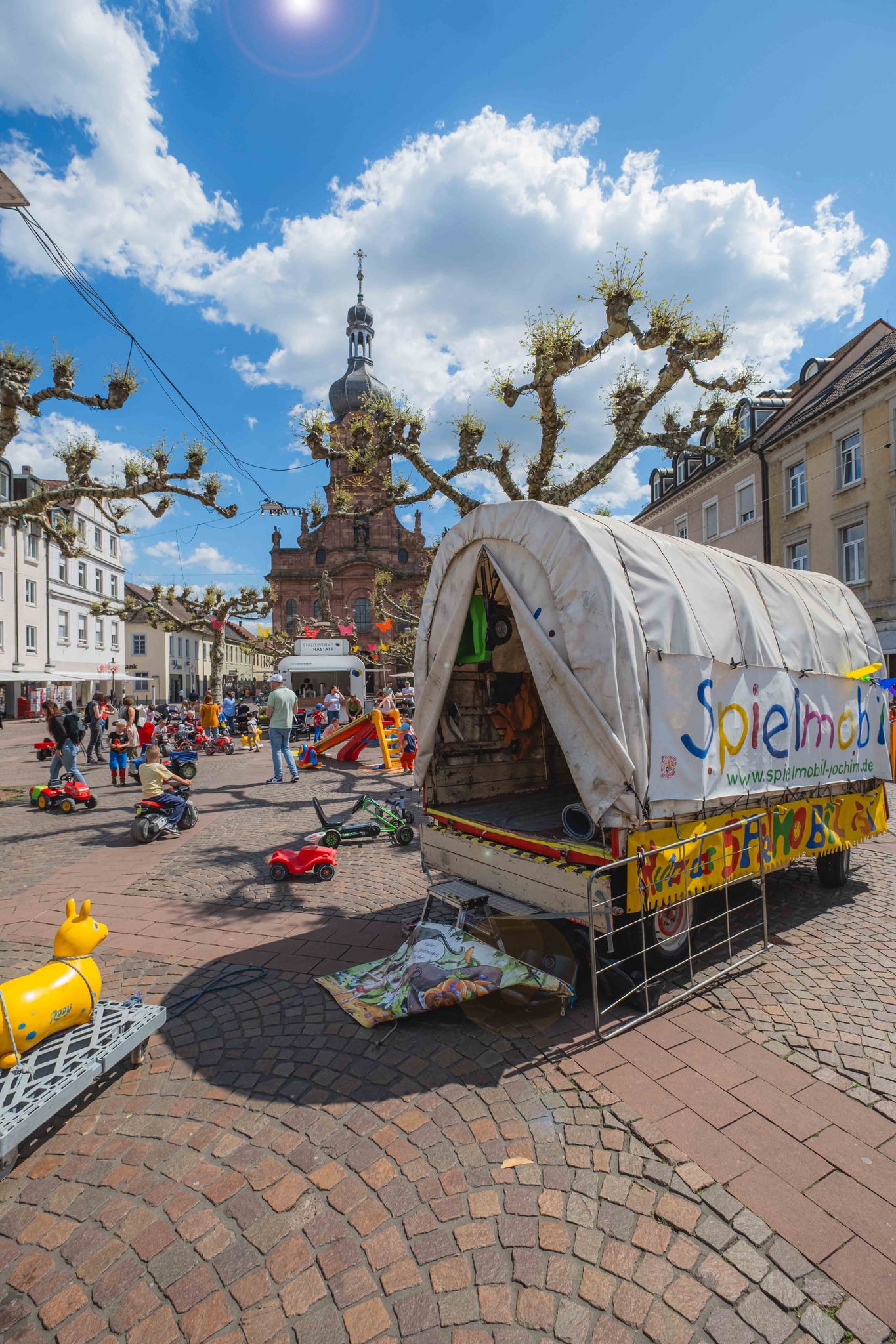 Dieser Alternativtext wurde KI-generiert: Ein heller Platz mit Gebäuden im Hintergrund und einer barocken Kirche. Im Vordergrund steht ein bunt dekorierter Wagen und es sind Menschen und Spielzeug zu sehen.