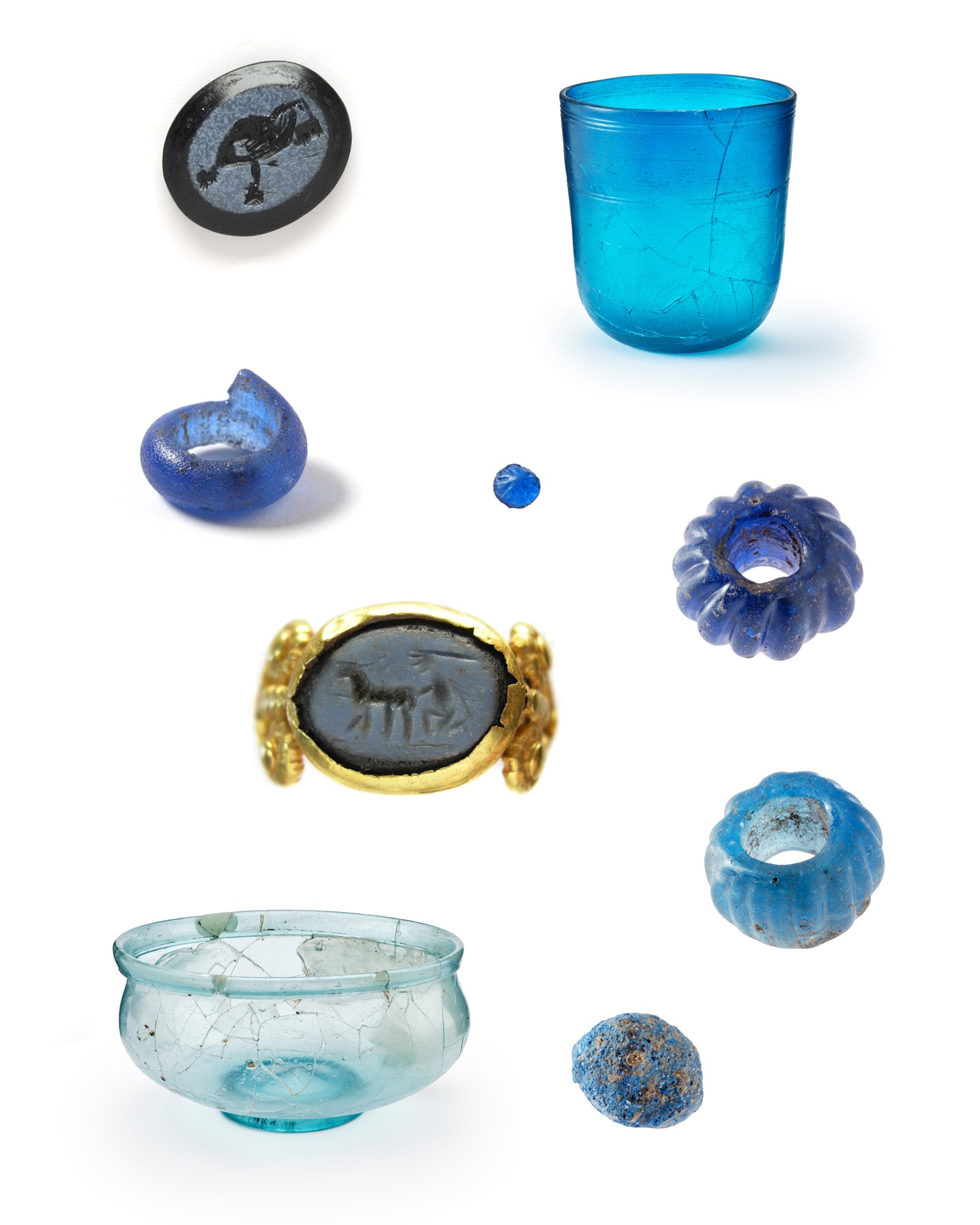 Eine Sammlung antiker Artefakte auf weißem Hintergrund. Dazu gehören ein blauer Glasbecher, eine helle Glasschale, mehrere blaue Glasperlen und -ringe sowie zwei Siegelsteine mit eingravierten Figuren. Ein goldener Ring mit einem dunklen Intaglio befindet sich in der Mitte. (KI-Alternativtext)