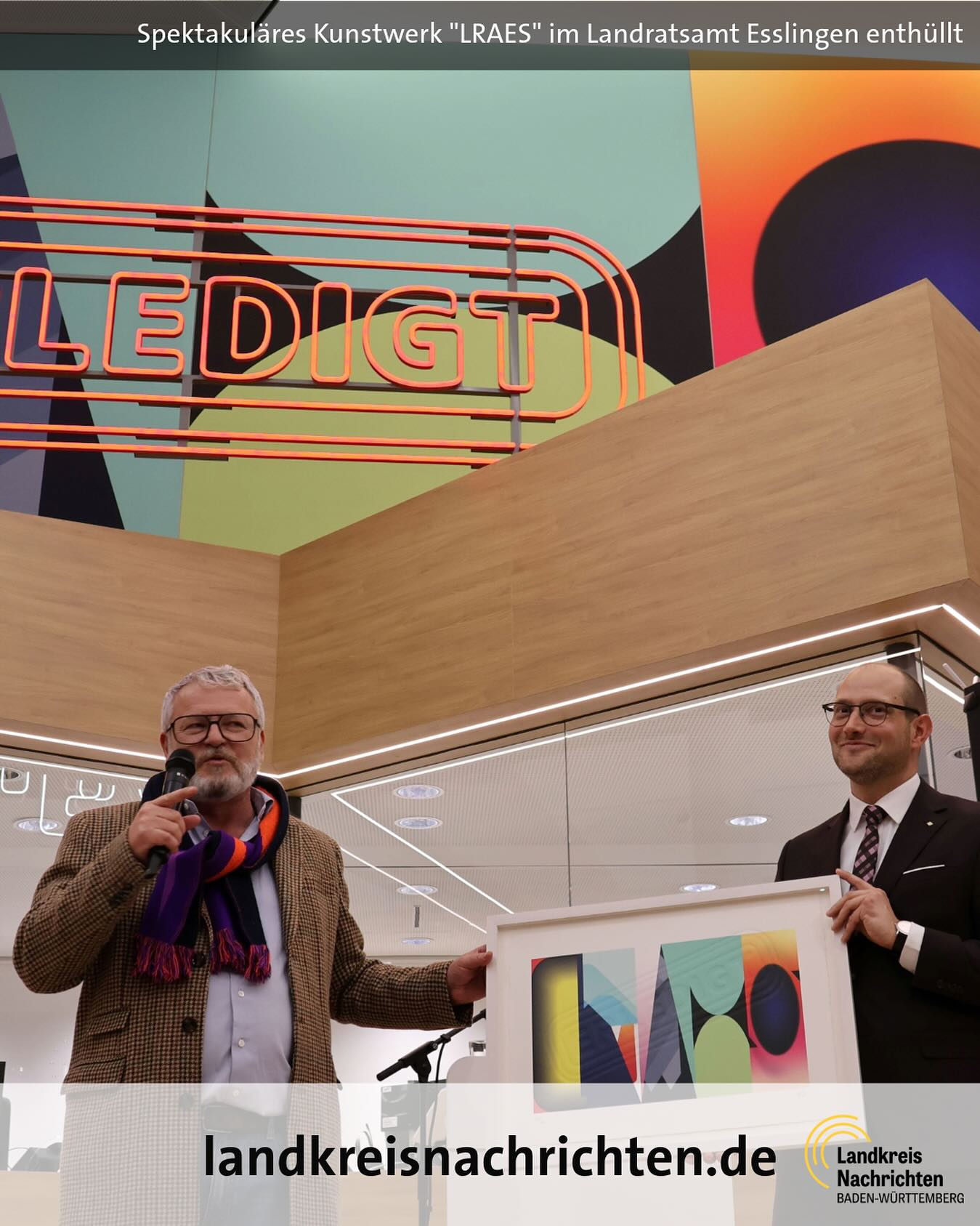 Dieser Alternativtext wurde KI-generiert: Ein farbenfrohes Kunstwerk ist im Hintergrund zu sehen. Zwei Personen stehen davor und halten etwas in den Händen, während sie fotografiert werden.