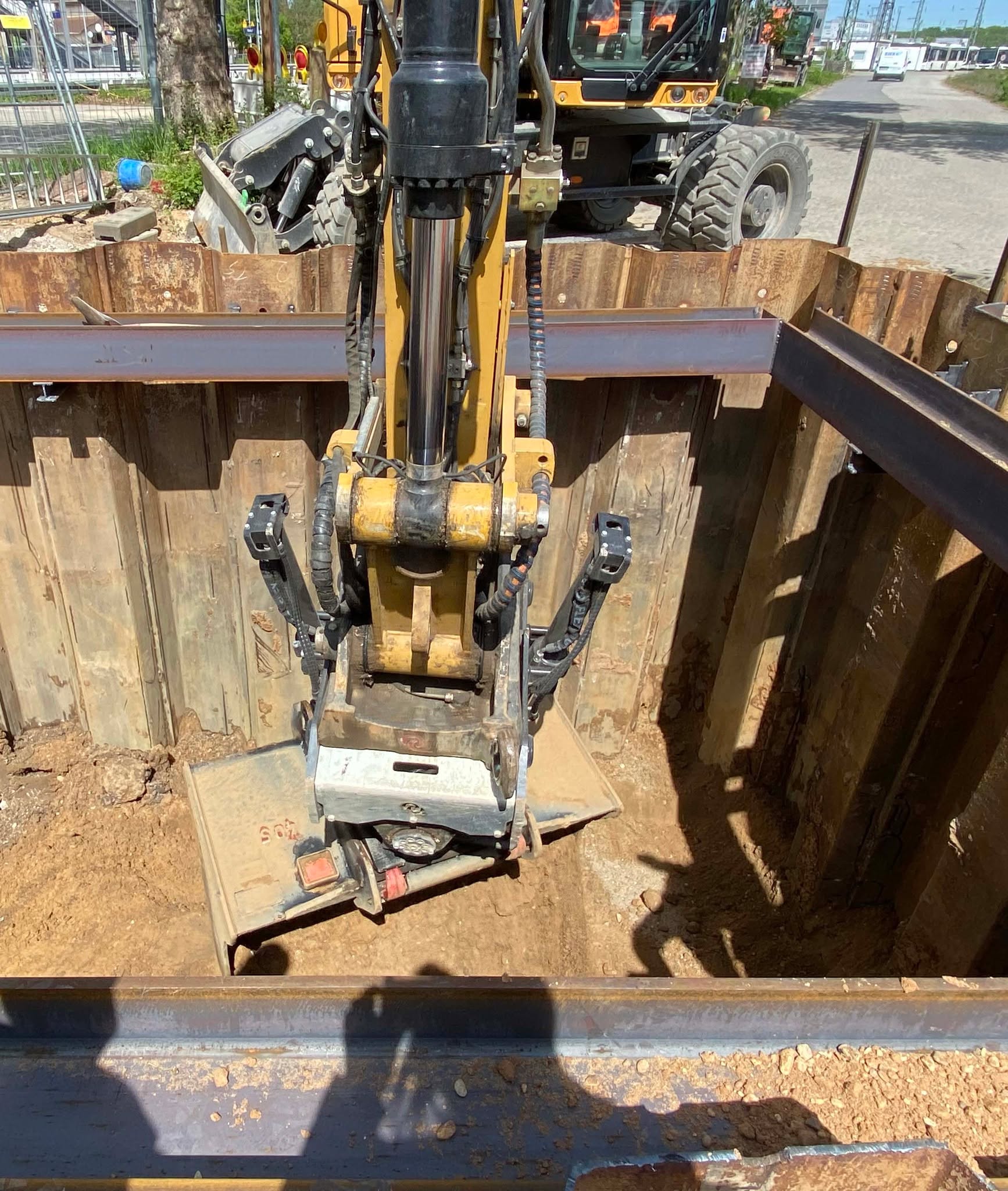 Dieser Alternativtext wurde KI-generiert: Eine Maschine mit einem großen Schaufelwerkzeug arbeitet in einer Baugrube. Die Umgebung zeigt eine Baustelle mit weiterer schwerer Technik und Baumaterialien.