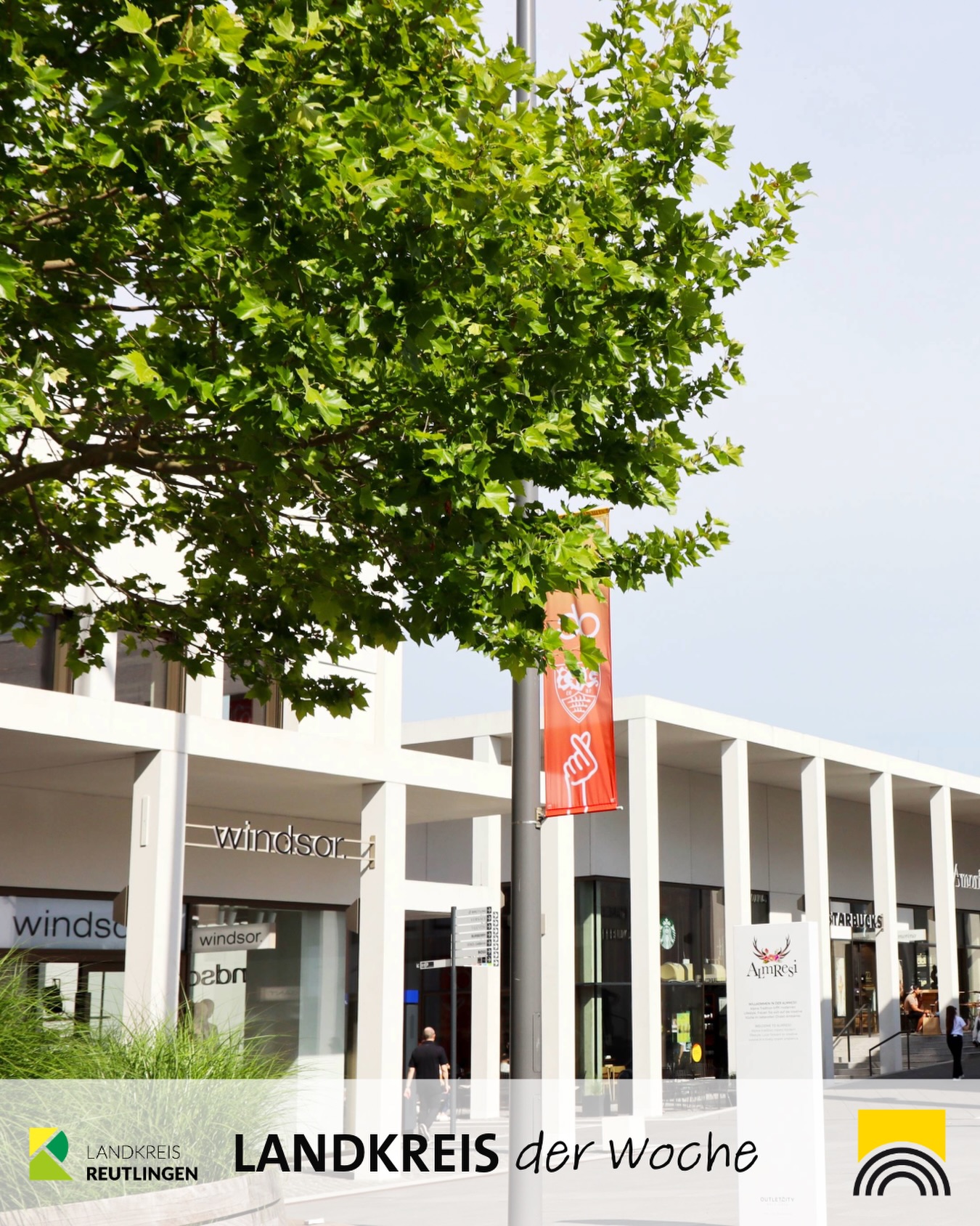 Dieser Alternativtext wurde KI-generiert: Ein helles Gebäude mit Geschäften ist teilweise von Bäumen verdeckt. Ein Banner mit einer grafischen Darstellung ist an der Fassade befestigt.