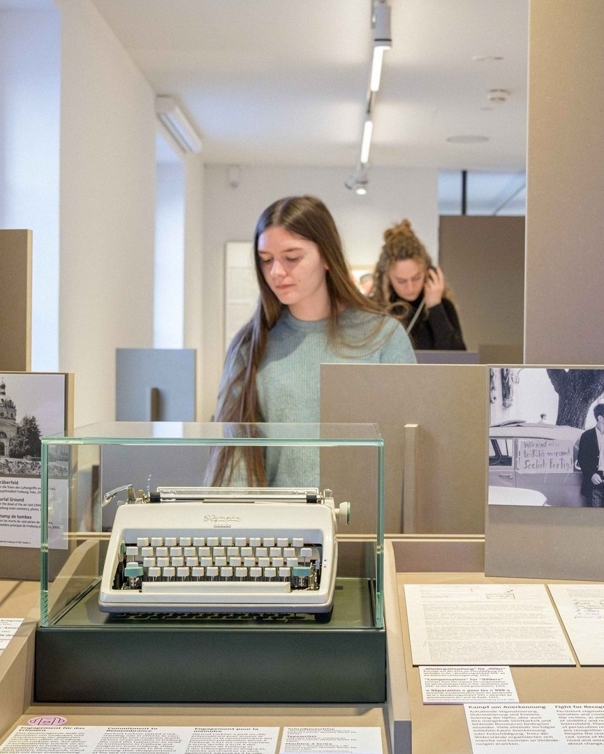 Im Vordergrund steht eine weiße Schreibmaschine in einer Glasschaustelle. Dahinter betrachtet eine Person mit langem Haar und blauem Pullover die Ausstellung. Im Hintergrund sind weitere Personen und Infotafeln, darunter eine mit der Überschrift „Kampf um Anerkennung“, zu erkennen. (KI-Alternativtext)