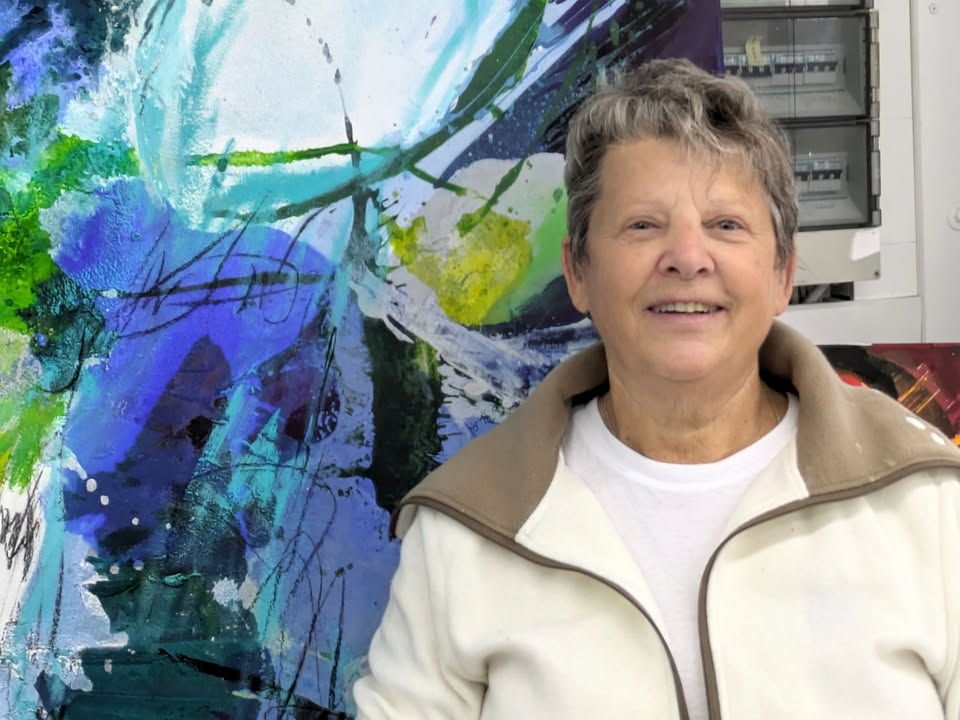 Dieser Alternativtext wurde KI-generiert: Eine Person lächelt in die Kamera, im Hintergrund ist ein abstraktes, farbenfrohes Gemälde zu sehen. Das Bild ist in Farbe gehalten.