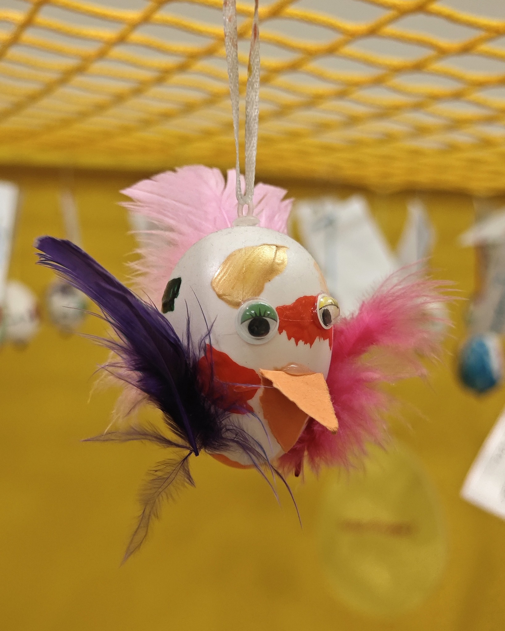 Dieser Alternativtext wurde KI-generiert: Eine bemalte, kugelförmige Figur mit Federn und Googly-Augen hängt an einer Schnur. Die Figur ist mit verschiedenen Farben und Mustern verziert.
