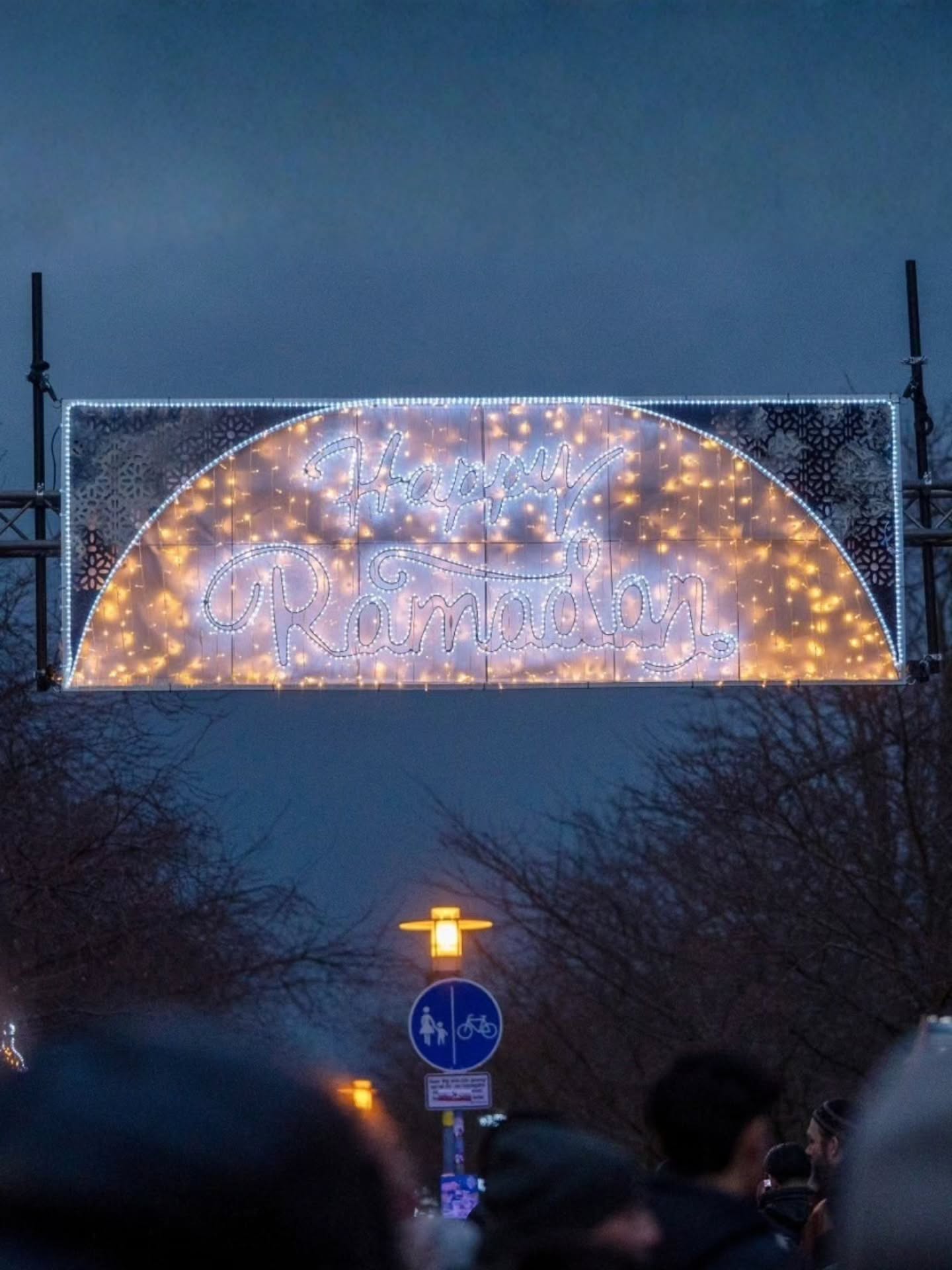 Dieser Alternativtext wurde KI-generiert: Ein beleuchteter Bogen mit der Aufschrift "Happy Ramadan" überspannt einen Weg. Im Vordergrund sind Silhouetten von Menschen und ein Verkehrsschild zu sehen.