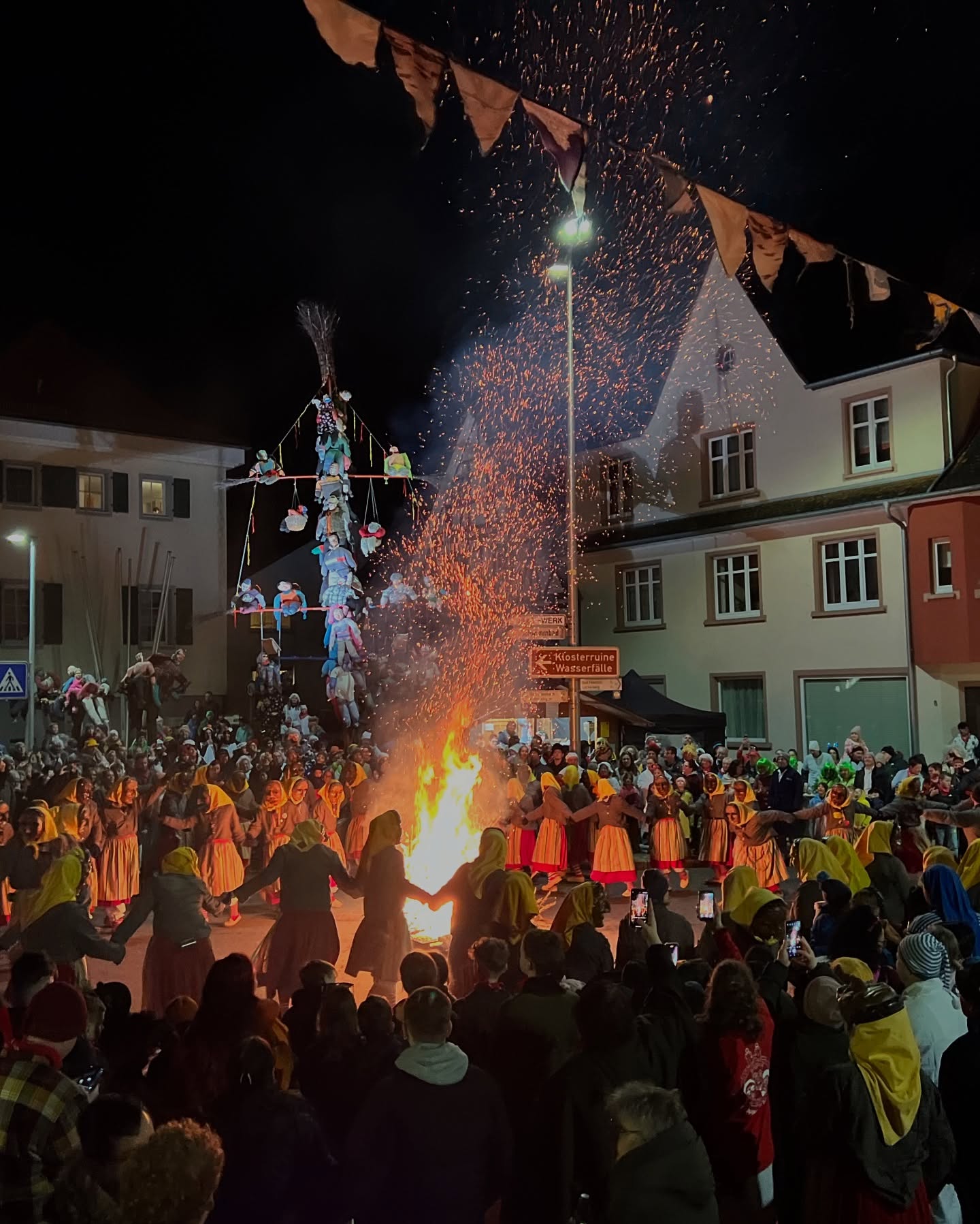 Eine Menschenmenge steht um ein Lagerfeuer. Über dem Feuer befindet sich eine dekorierten Konstruktion.