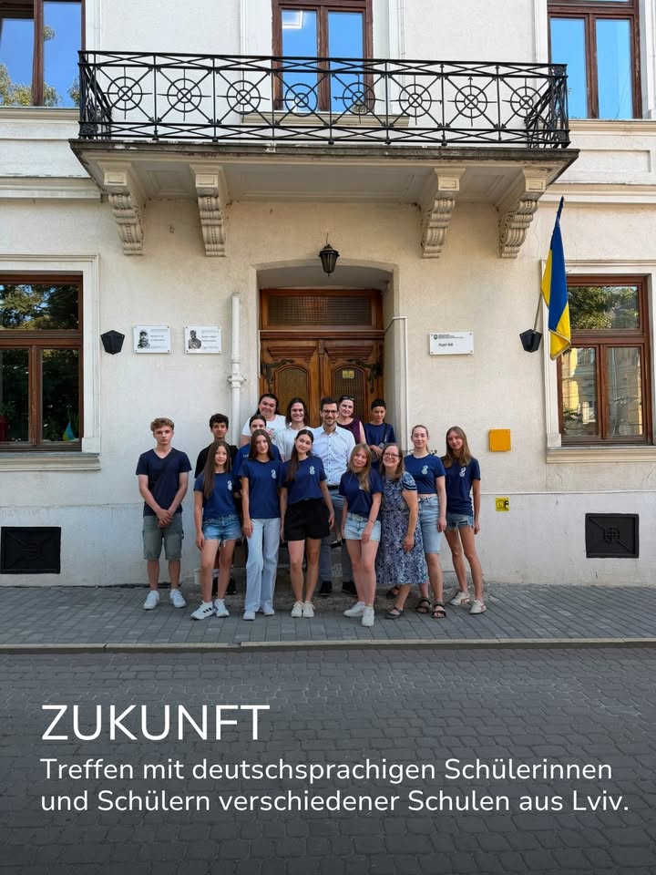 Eine Gruppe von Personen, viele davon in blauen T-Shirts, steht vor dem Eingang eines Gebäudes mit ukrainischer Flagge. Im unteren Bildbereich steht der weiße Text: „ZUKUNFT Treffen mit deutschsprachigen Schülerinnen und Schülern verschiedener Schulen aus Lviv.“ (KI-Alternativtext)