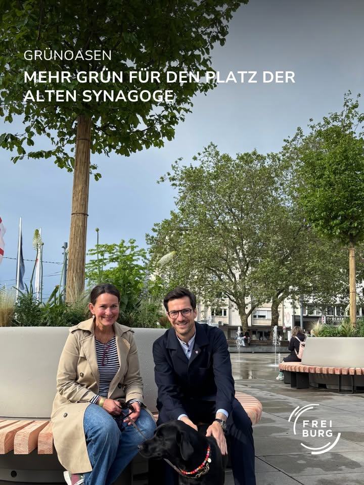 Dieser Alternativtext wurde KI-generiert: Ein Foto zeigt zwei Personen und ein Tier vor Bäumen und Gebäuden. Im Vordergrund befindet sich eine Bank und ein Logo.