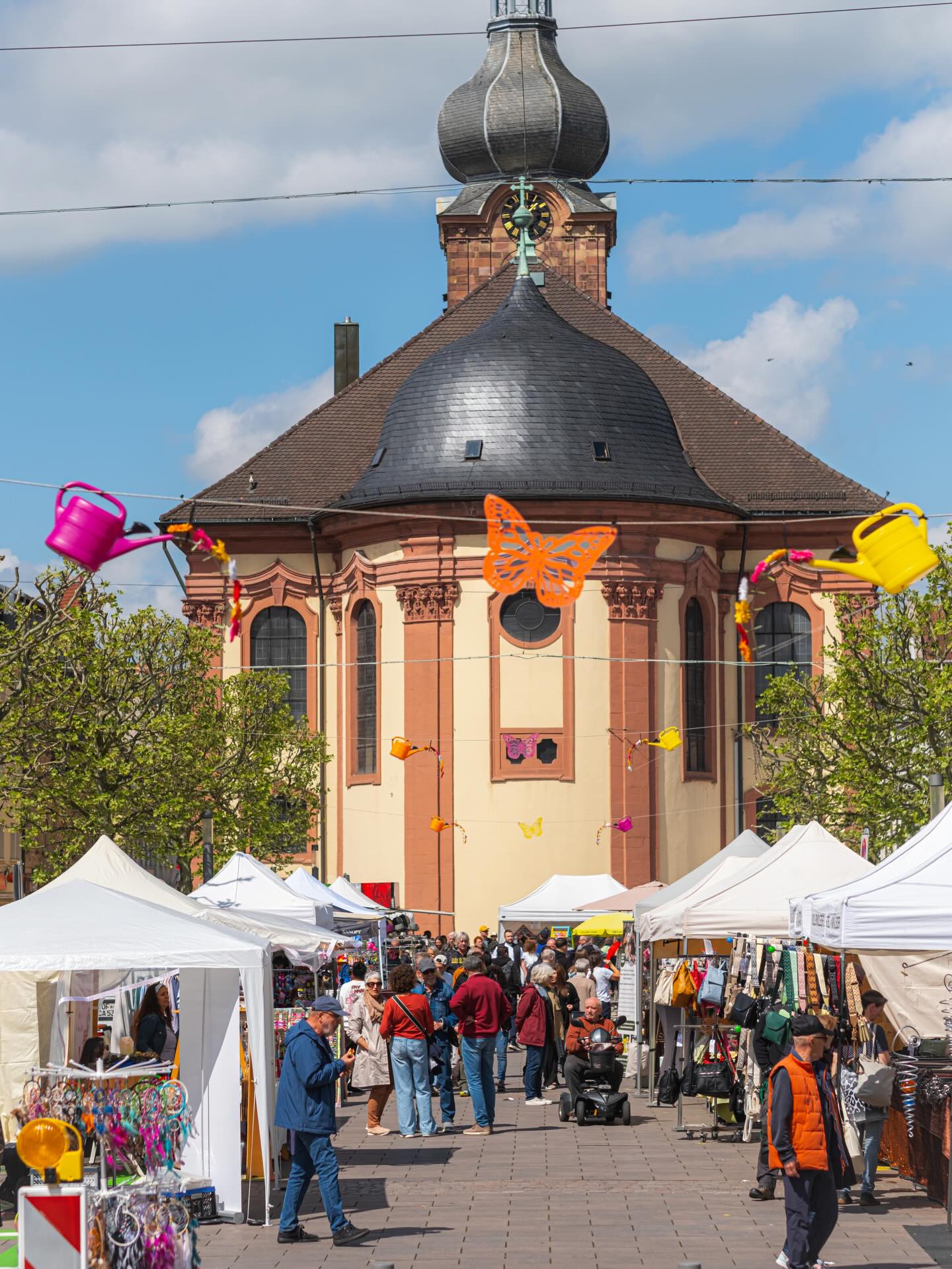 Dieser Alternativtext wurde KI-generiert: Ein rosafarbenes Gebäude mit einem hohen, geschwungenen Dach und einer Uhr. Vor dem Gebäude befindet sich ein belebter Markt mit Ständen und Menschen.