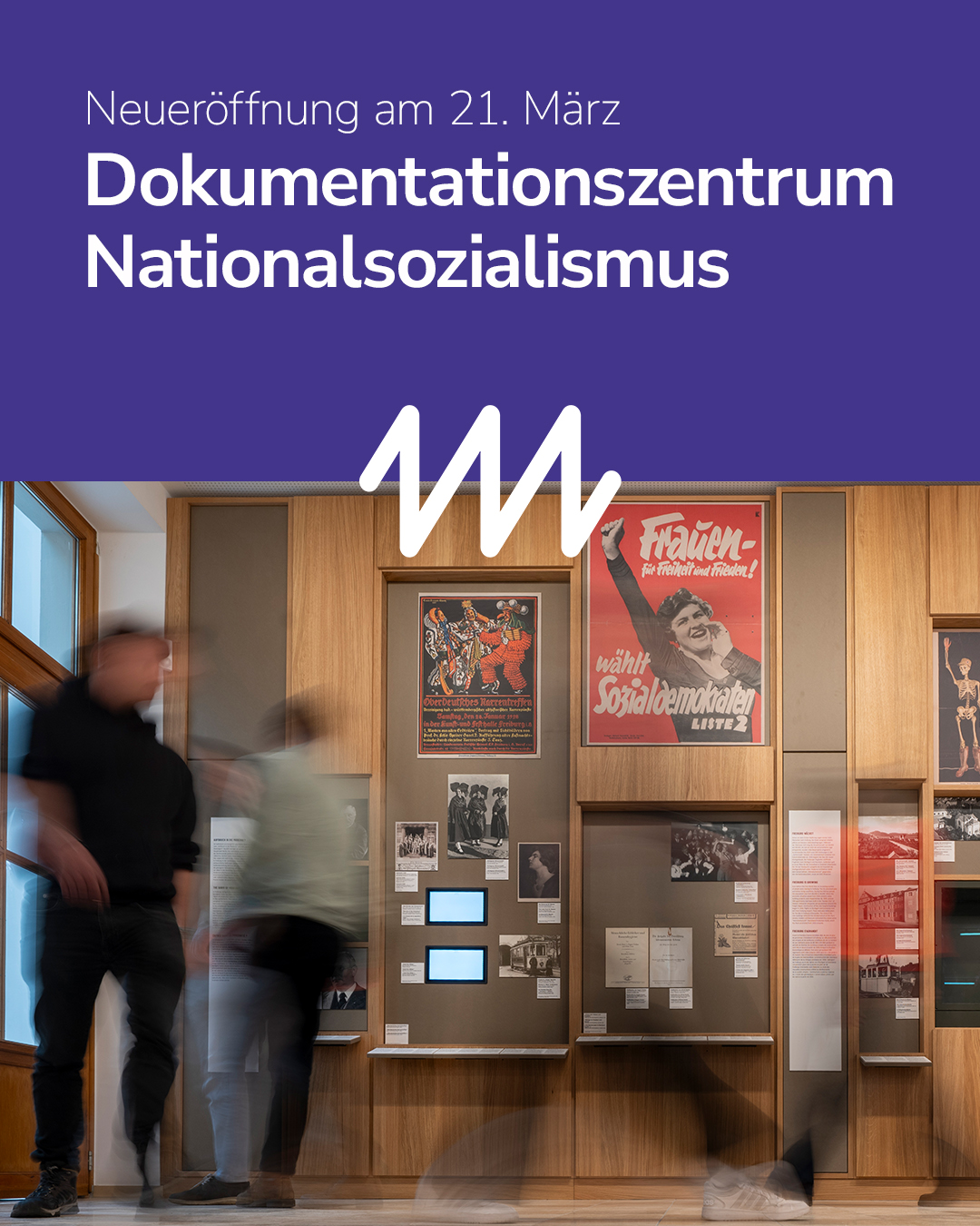 Bild eines modernen Ausstellungsraums mit historischen Plakaten und Fotografien. Mehrere Personen sind im Raum zu sehen, einige sind unscharf dargestellt.