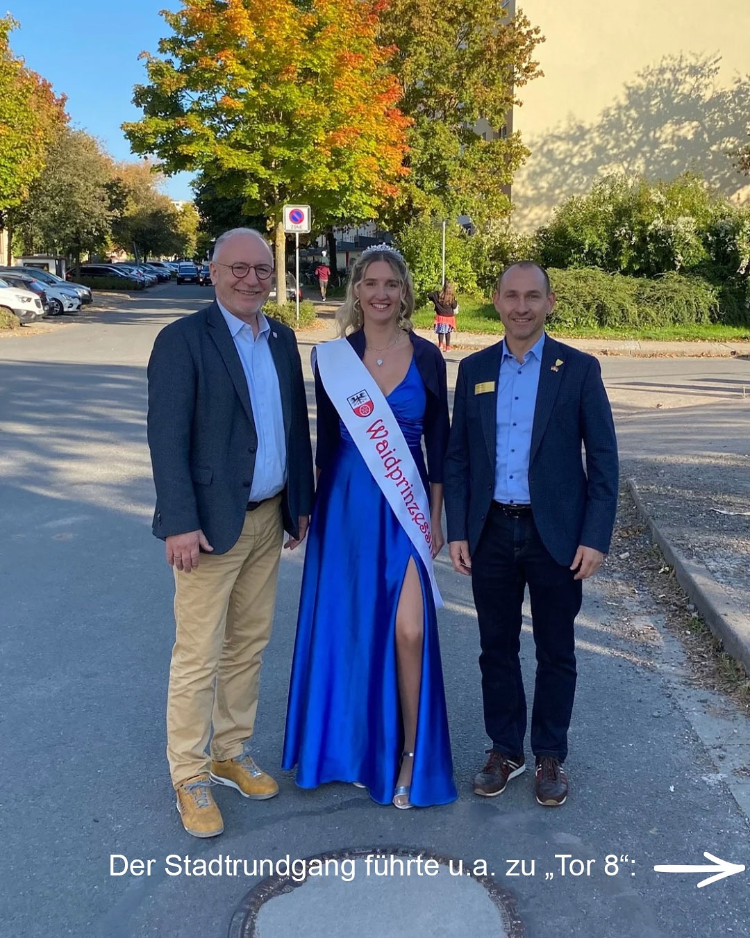 Dieser Alternativtext wurde KI-generiert: Drei Personen stehen auf einem Bürgersteig vor Bäumen und einer Straße. Eine Person trägt ein blaues Kleid mit einem roten Band und eine Tiara.