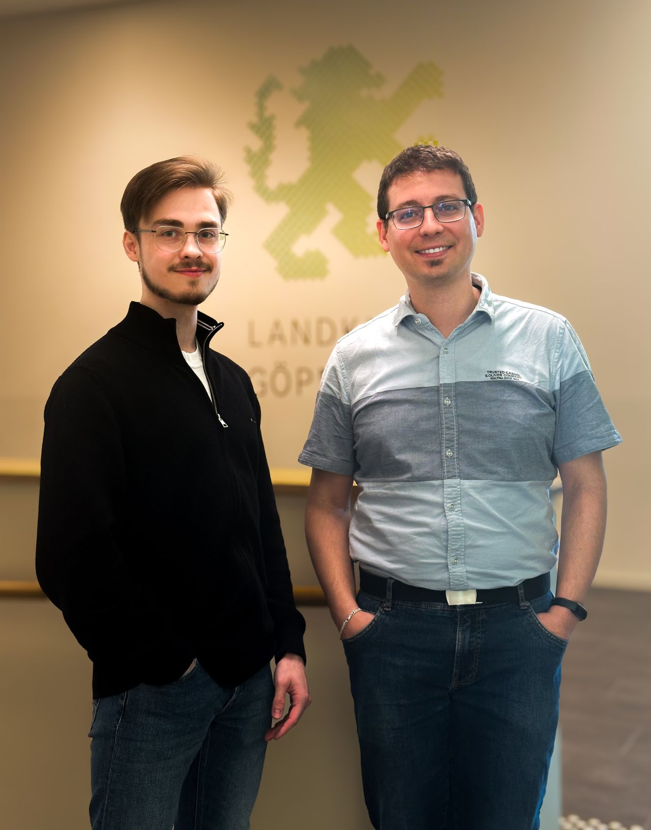 Dieser Alternativtext wurde KI-generiert: Zwei Personen stehen nebeneinander vor einem hellen Hintergrund mit einem Wappen. Das Bild ist farbig und zeigt die beiden Personen von der Brust aufwärts.