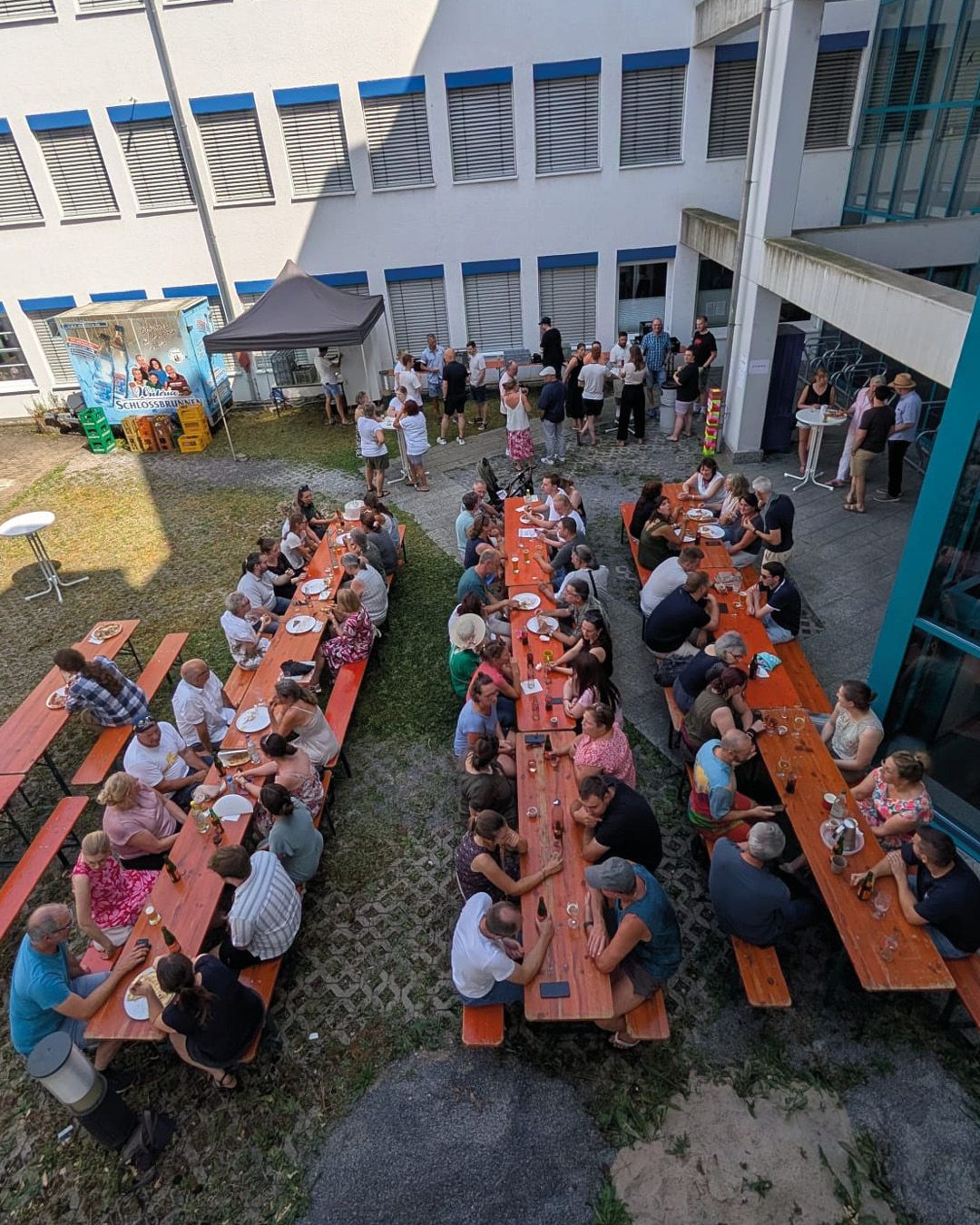 Viele Personen sitzen an langen Holztischen auf einer Wiese und einem gepflasterten Hof, wo sie essen und trinken. Im Hintergrund stehen weitere Personen in Gruppen und unterhalten sich vor einem weißen Gebäude mit blauen Fensterrahmen. Links im Bild steht ein Zelt und ein großes Schild mit der Aufschrift „Schlossbrünner“. (KI-Alternativtext)