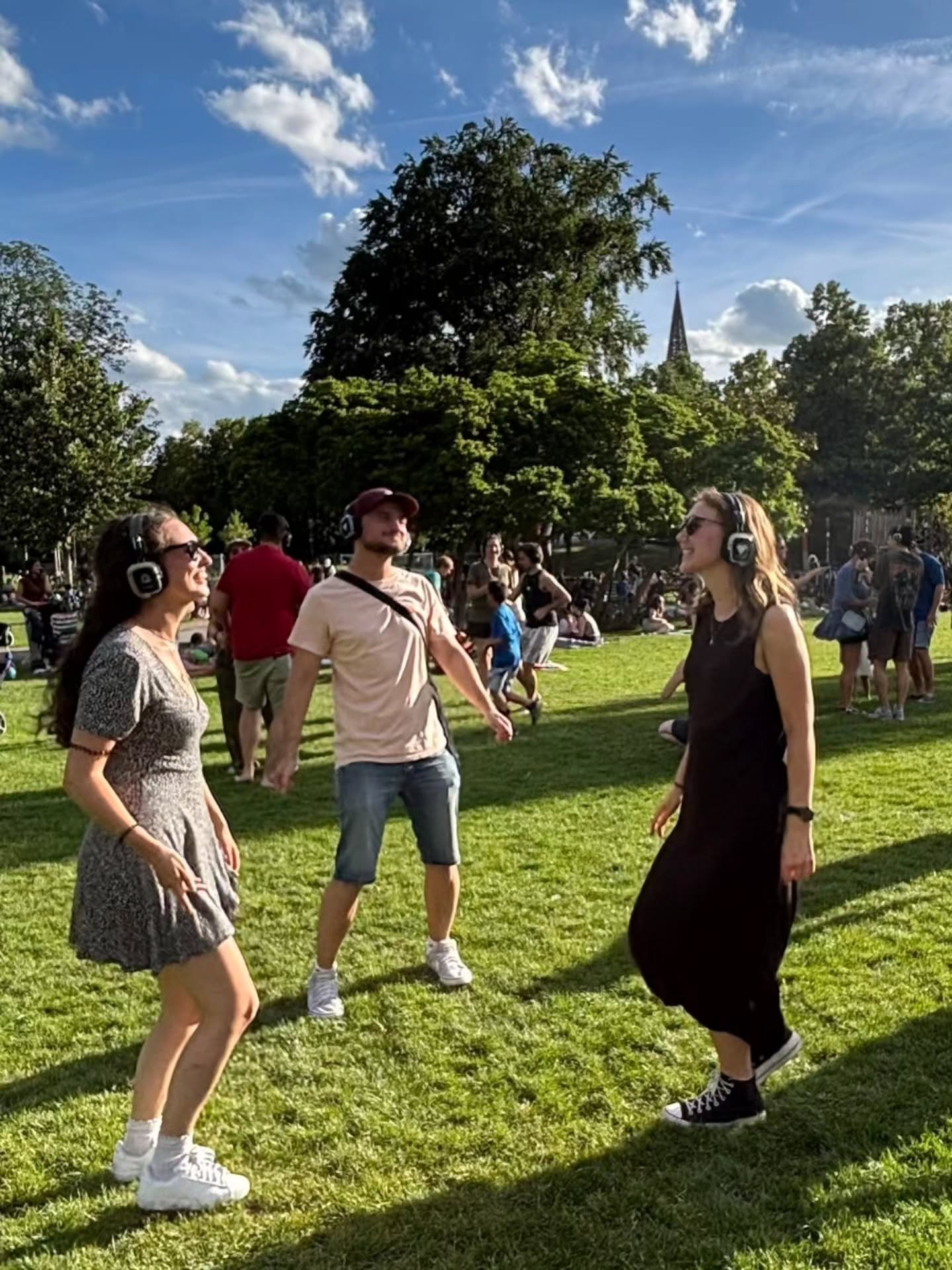 Drei Personen tanzen auf einer Wiese im Freien, alle tragen kabellose Kopfhörer. Im Hintergrund sind weitere Menschen, Bäume und ein Kirchturm unter blauem Himmel mit Wolken zu sehen. (KI-Alternativtext)
