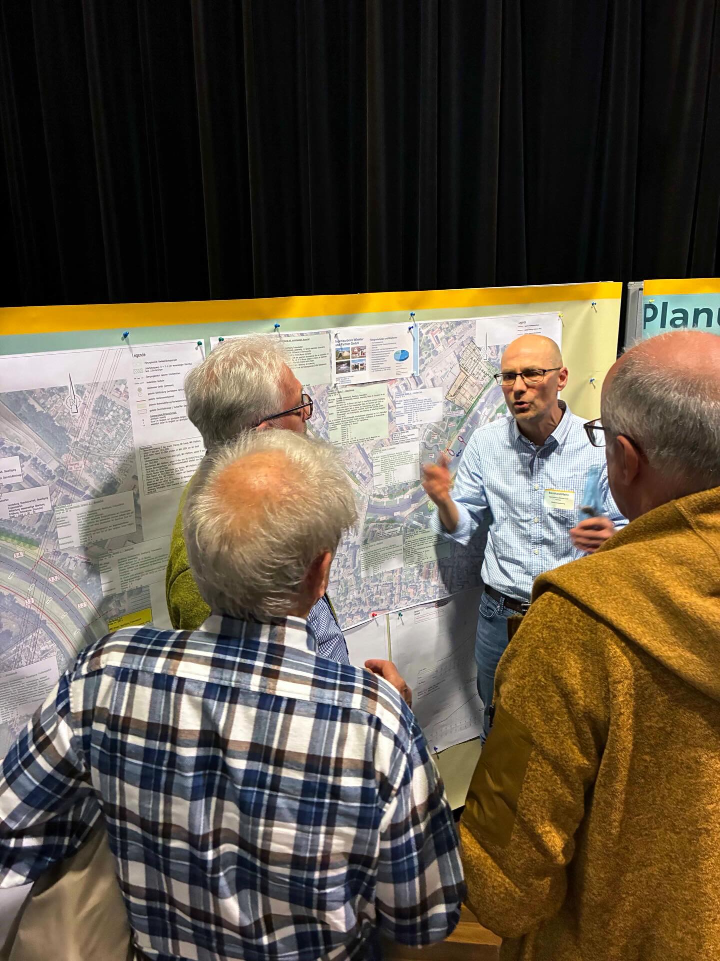 Dieser Alternativtext wurde KI-generiert: Mehrere Personen betrachten einen Plan, der an einer Stellwand hängt. Eine Person deutet auf den Plan, während die anderen zuhören.