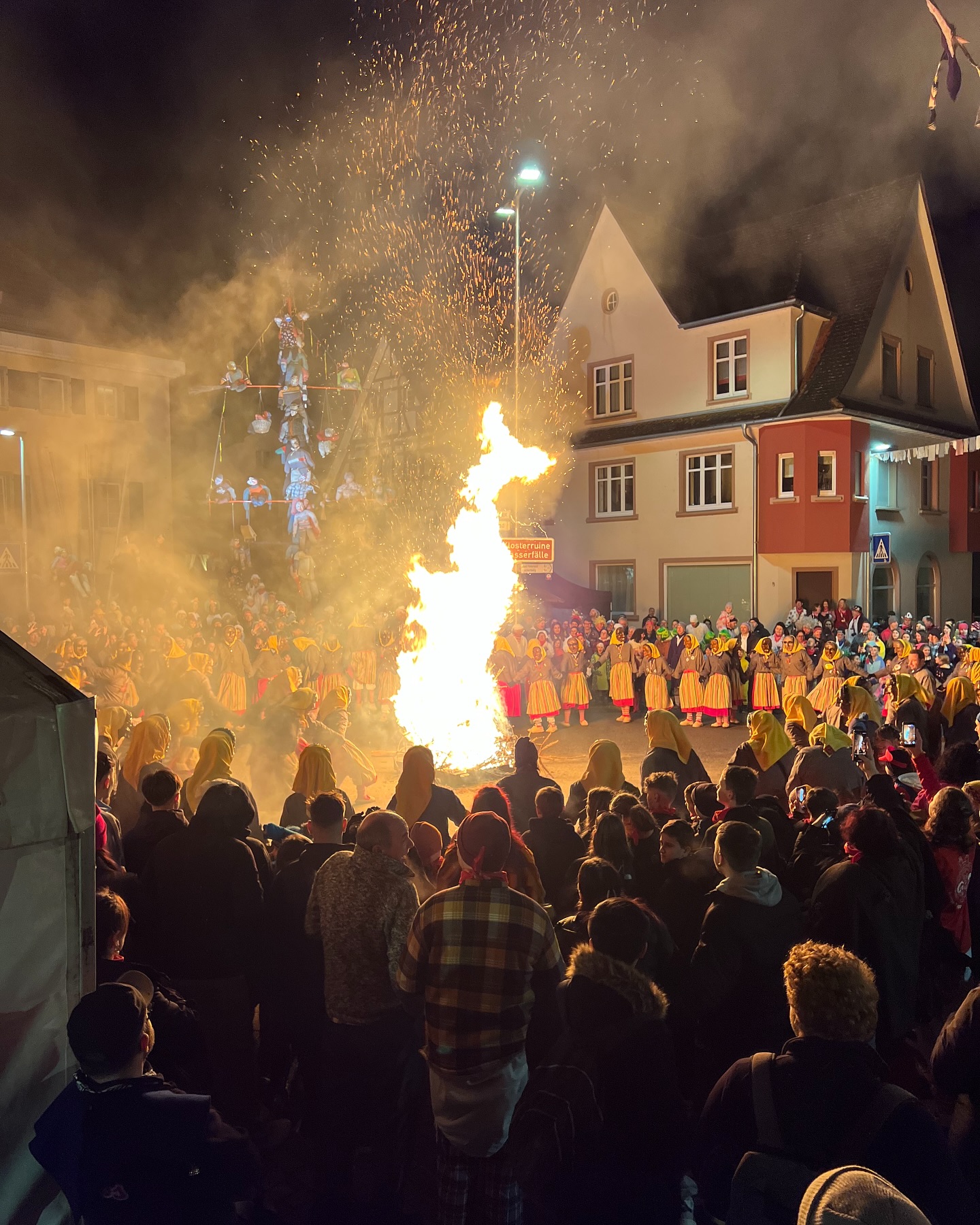 A crowd gathers around a large bonfire at night. In the background, there are figures on a tall structure and people in yellow robes holding hands.