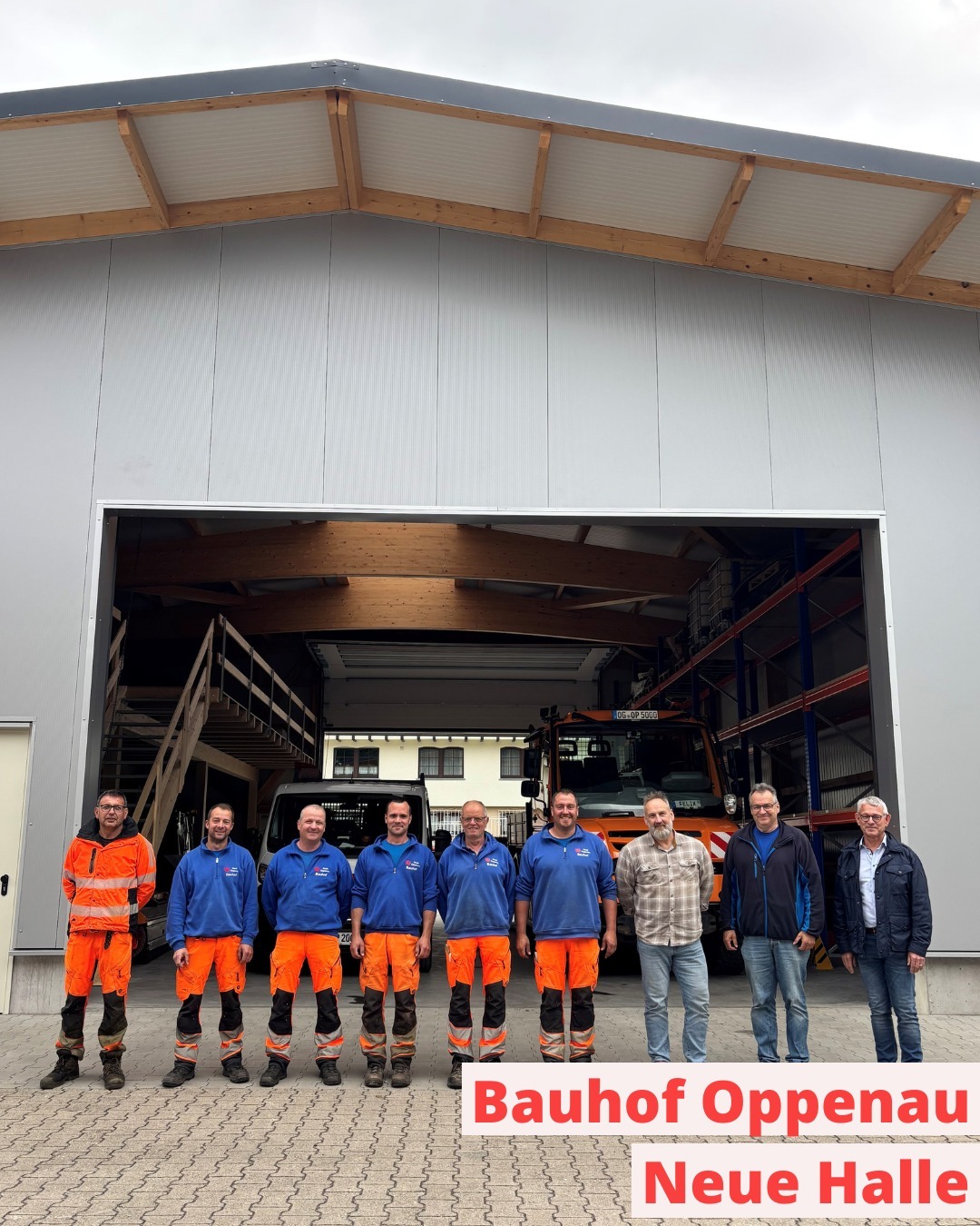 Acht Personen stehen vor einer großen Halle mit geöffneter Tür, dahinter sind ein Fahrzeug und Regale zu sehen. Einige tragen orangefarbene Arbeitskleidung, andere Alltagskleidung. Unten steht: „Bauhof Oppenau Neue Halle“. (KI-Alternativtext)