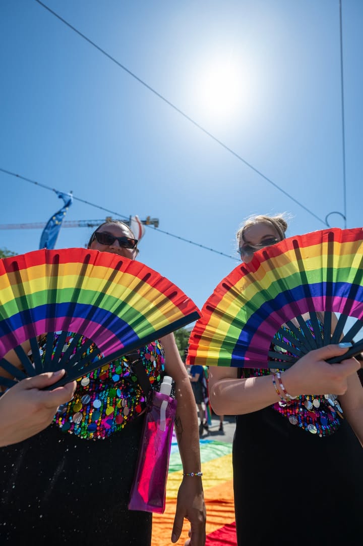 Zwei Personen halten bunte Regenbogenfächer vor sich, tragen glitzernde Oberteile und Sonnenbrillen, im Hintergrund strahlender Sonnenschein und ein blauer Himmel mit Stromleitungen. (KI-Alternativtext)