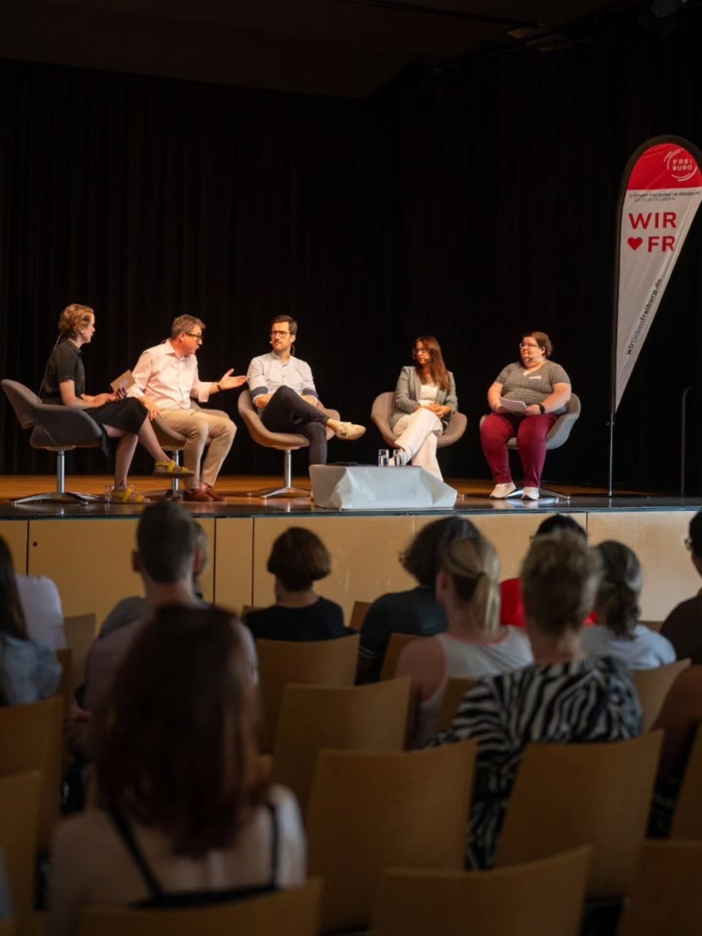 Fünf Personen sitzen auf Stühlen auf einer Bühne vor einem Publikum und führen eine Diskussion. Rechts im Hintergrund steht ein Werbebanner mit der Aufschrift „WIR ♥ FR“ und „FREIBURG“. Im Vordergrund sind die Köpfe von Zuschauern auf Stühlen zu sehen. (KI-Alternativtext)