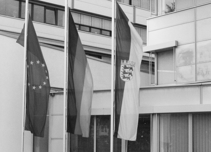 Dieser Alternativtext wurde KI-generiert: Schwarz-Weiß-Foto von drei Flaggen vor einem Gebäude. Die Flaggen zeigen unterschiedliche Muster und Symbole.